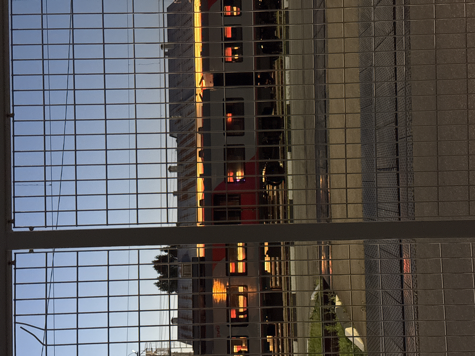 A train with illuminated windows is visible behind a wire fence in Moscow, Russia. The train appears to be gray and red, with bright orange light reflecting in its windows. The sky above is a clear blue, and the scene suggests it is either dawn or dusk. In the background, there are buildings with metal roofs and some trees. The foreground is dominated by the wire fence and a paved area. The overall impression is of a railway station or a similar industrial area in the city.