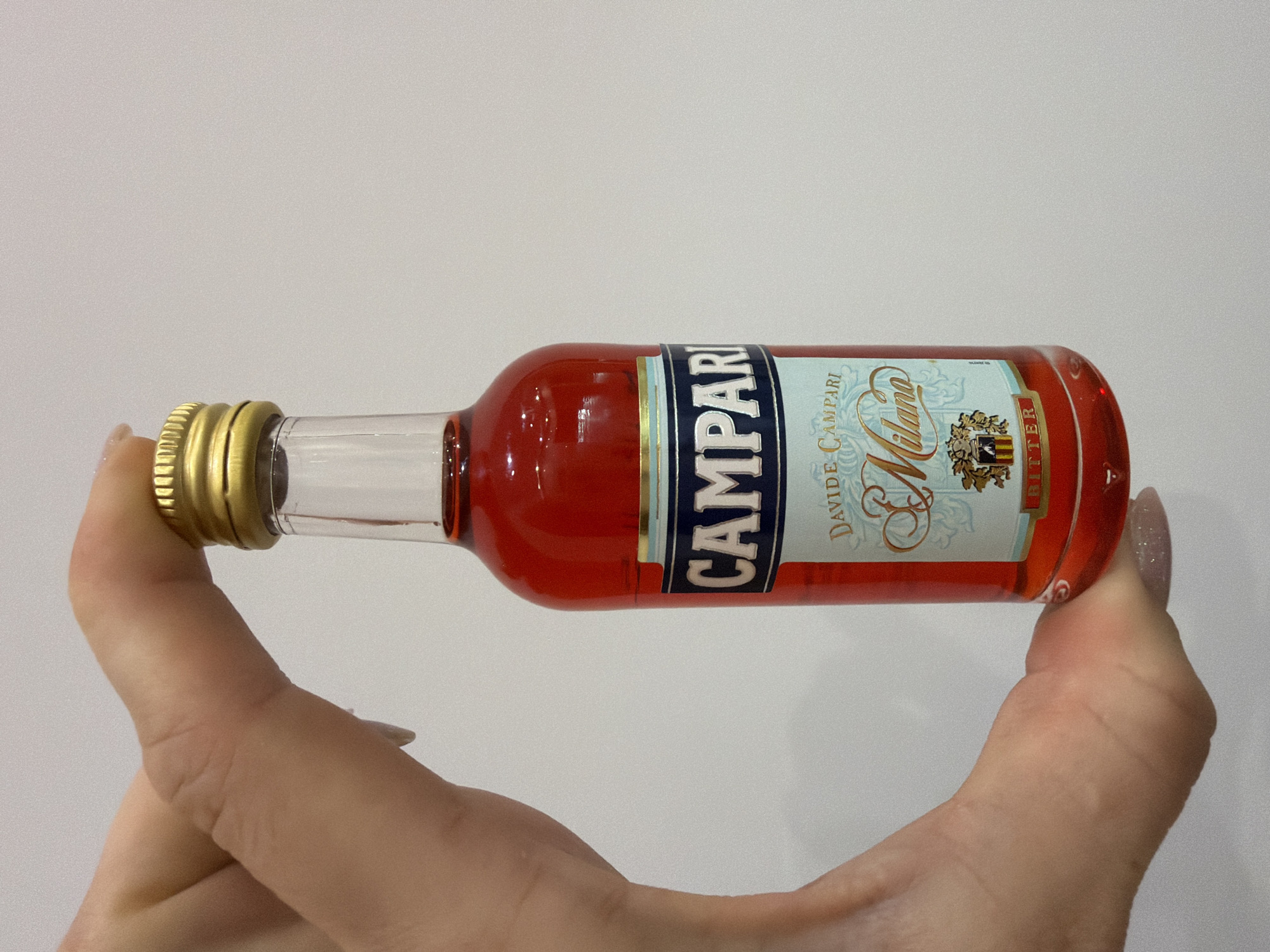 A person's hand with manicured nails is holding a small, clear glass bottle containing a red liquid. The bottle has a gold cap and a label that reads "CAMPARI," "DAVIDE. CAMPARI," "Milano," and "BITTER." The background is a plain, light-colored wall. The scene is indoors.