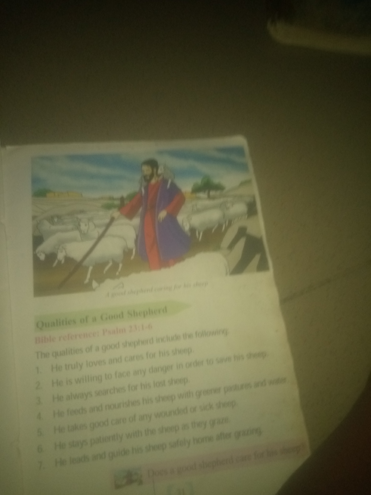 The image displays a page from a book with an illustration of a shepherd tending to his flock. The illustration depicts a man in robes, holding a staff, with several sheep around him in a pastoral setting. Below the illustration, there is text that reads "Qualities of a Good Shepherd" and "Bible reference: Psalm 23:1-6." A numbered list follows, detailing the qualities of a good shepherd, such as loving and caring for his sheep, facing danger, searching for lost sheep, feeding and nourishing them, and leading them home. The bottom of the page has a small graphic and the text "Does a good shepherd care for his sheep?". The image is blurry and angled, making it difficult to discern fine details. There is no information within the image or its context that indicates its location in Jauro Yinu, Nigeria.
