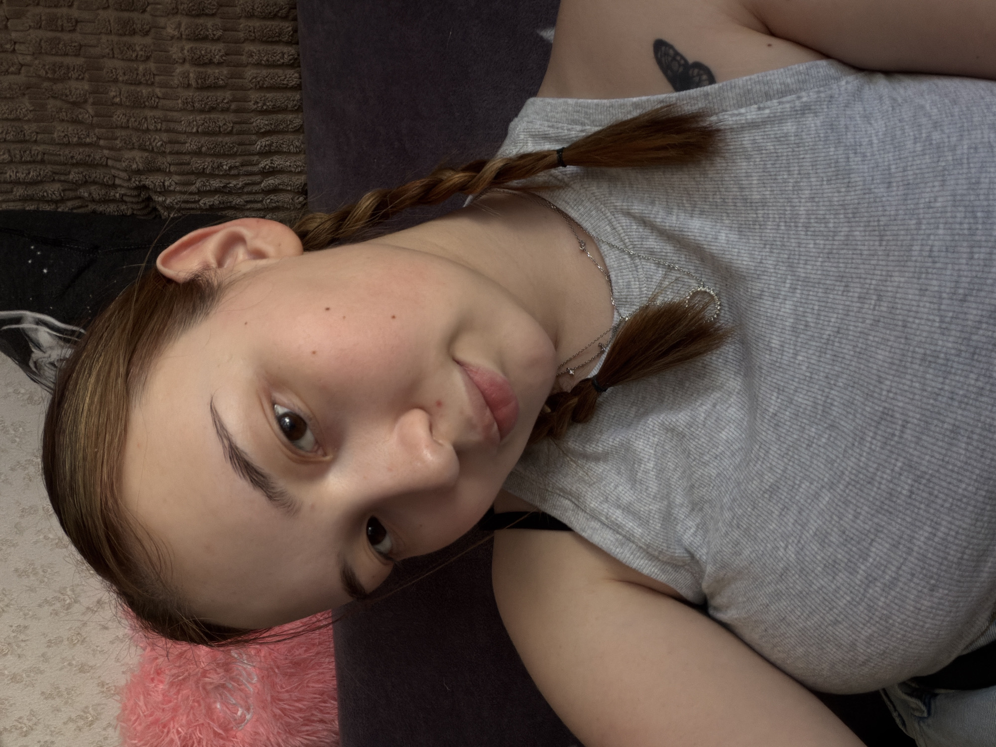 A young woman lies on a bed in an indoor setting. She has light brown hair styled in two braids and wears a light grey ribbed tank top. A silver necklace adorns her neck, and a small black butterfly tattoo is visible on her upper left arm. The background includes a textured brown surface and a dark grey pillow or cushion. A pink fuzzy object and a patterned surface are visible in the lower left corner. The location in the city of Chaikovsky, Russia, cannot be determined from the provided image.