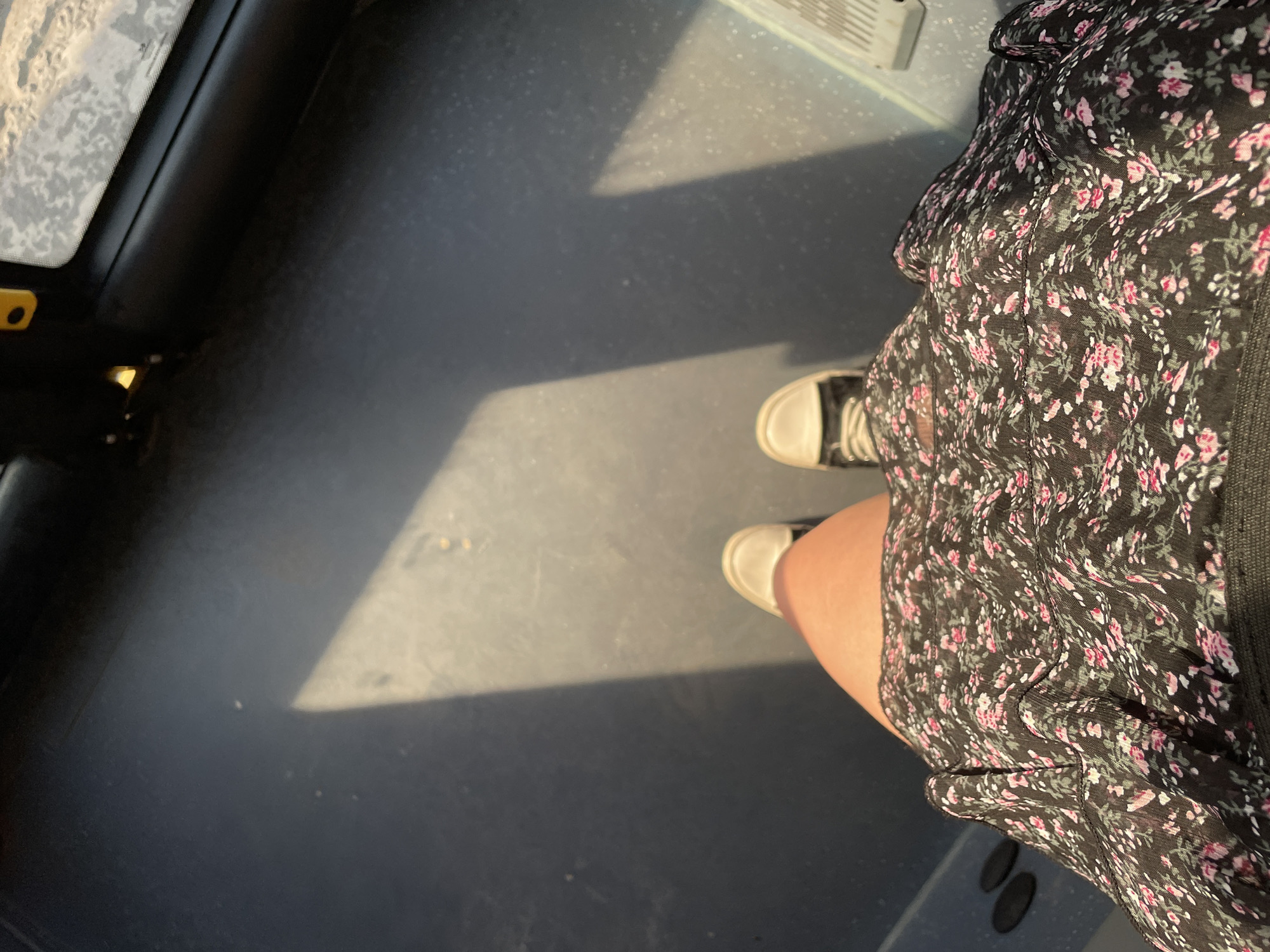 A low-angle perspective shot shows a person's legs from the knees down, wearing a floral print dress and sneakers, standing on a dark grey floor. Sunlight casts a sharp, rectangular shadow across the floor from the left side of the frame. The floor appears to be inside a vehicle, potentially a bus or train, with a hint of a window with frosted glass visible in the upper right corner, and a yellow grab handle in the upper left. The overall environment is dimly lit, with the sunlight creating a distinct contrast.