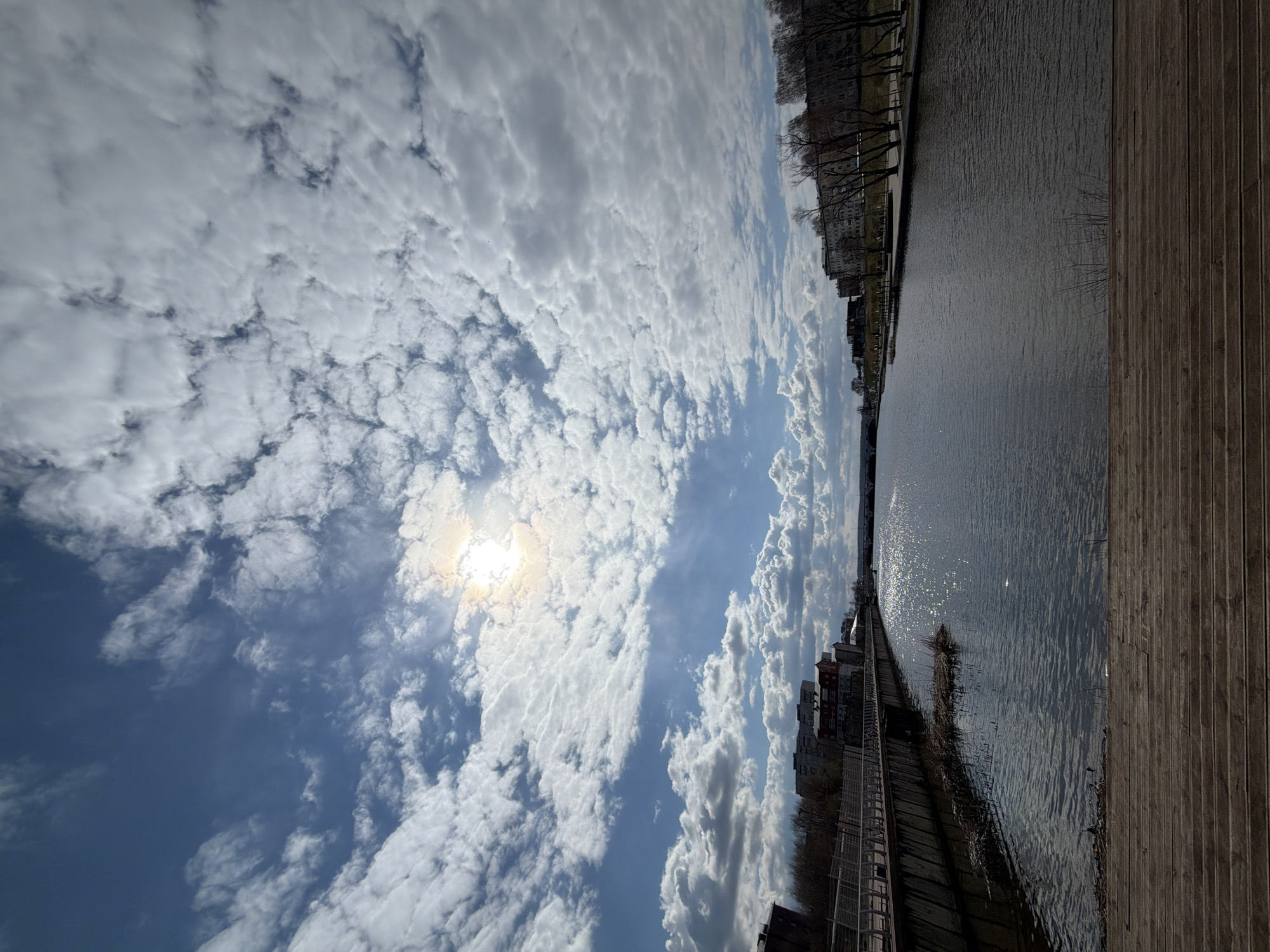 The image depicts a view from a waterfront in Votkinsk, Russia. The foreground features a wooden boardwalk running along the right side of the frame. To the left of the boardwalk is a body of water, likely a section of the Votkinsk Reservoir, with sunlight reflecting on its surface. On the far side of the water are buildings and trees lining the shore. The sky is filled with a variety of clouds, with the sun visible through a thin area of cloud cover.