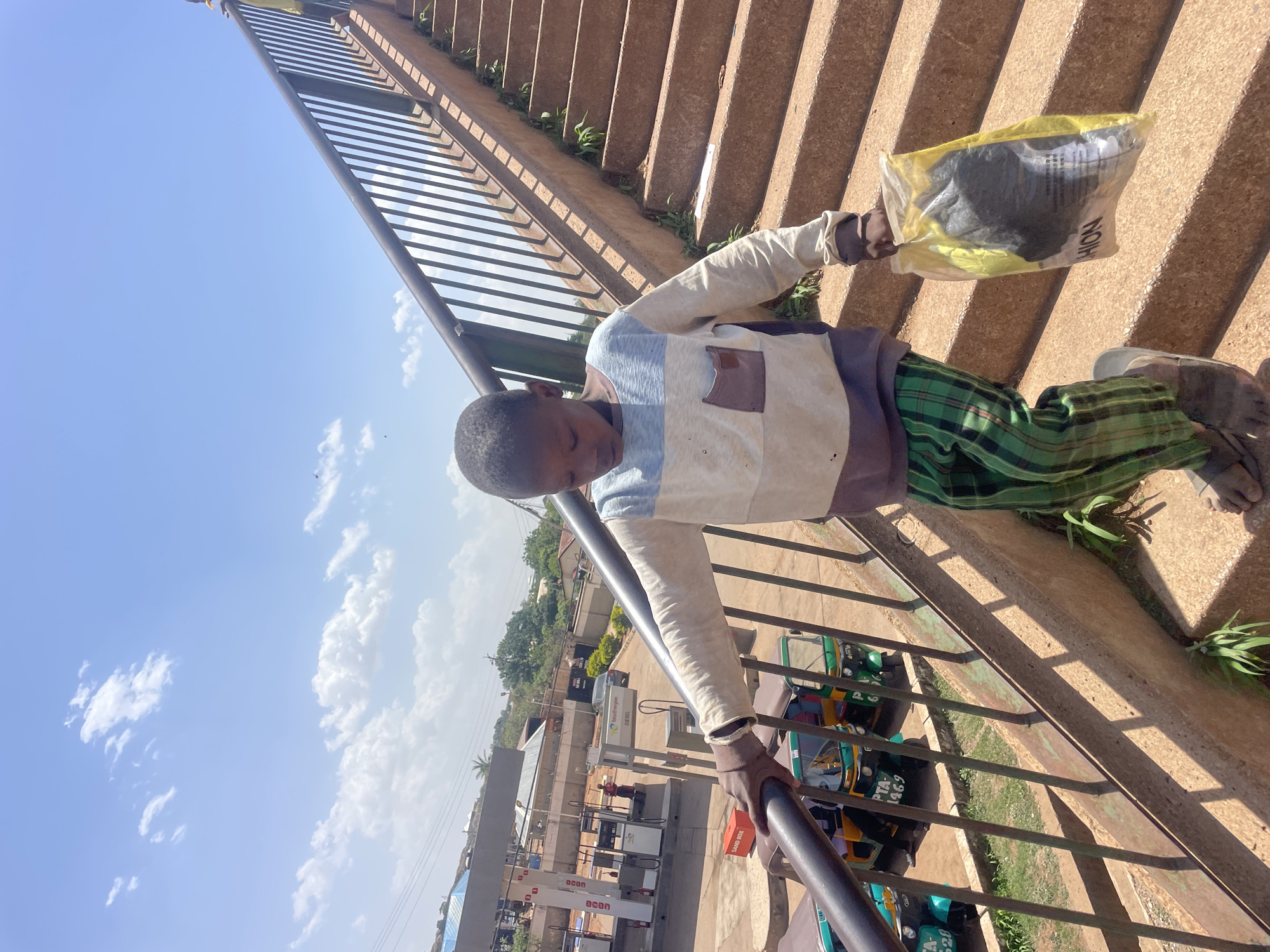 A person stands on a pedestrian bridge in Gura Top, Nigeria, holding a plastic bag. They are positioned on a series of concrete steps with a metal railing. The individual is wearing a long-sleeved shirt with horizontal stripes and green patterned pants. Below the bridge, a gas station and several auto rickshaws (tuk-tuks) are visible. The sky is blue with scattered clouds.