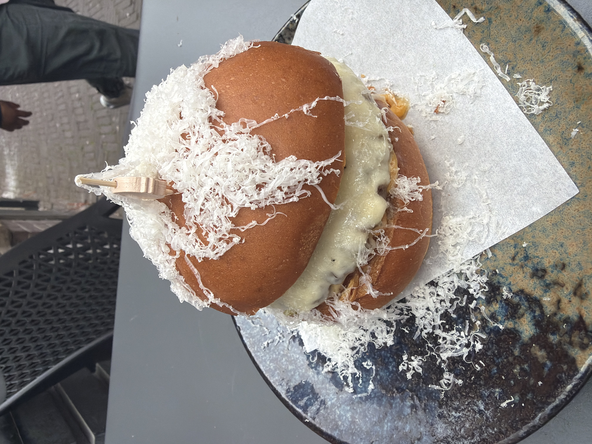 A close-up, eye-level shot shows a gourmet burger with a generous amount of shredded cheese on top, presented on a speckled blue and brown plate. The burger, with its golden-brown bun and melted cheese patty, is partially obscuring a white paper liner. The plate rests on a dark grey surface, possibly an outdoor table. In the blurred background, a dark metal grate structure is visible to the left, and to the right, a person in dark trousers and a jacket is walking on a cobblestone path. The scene suggests an outdoor dining setting in a city.