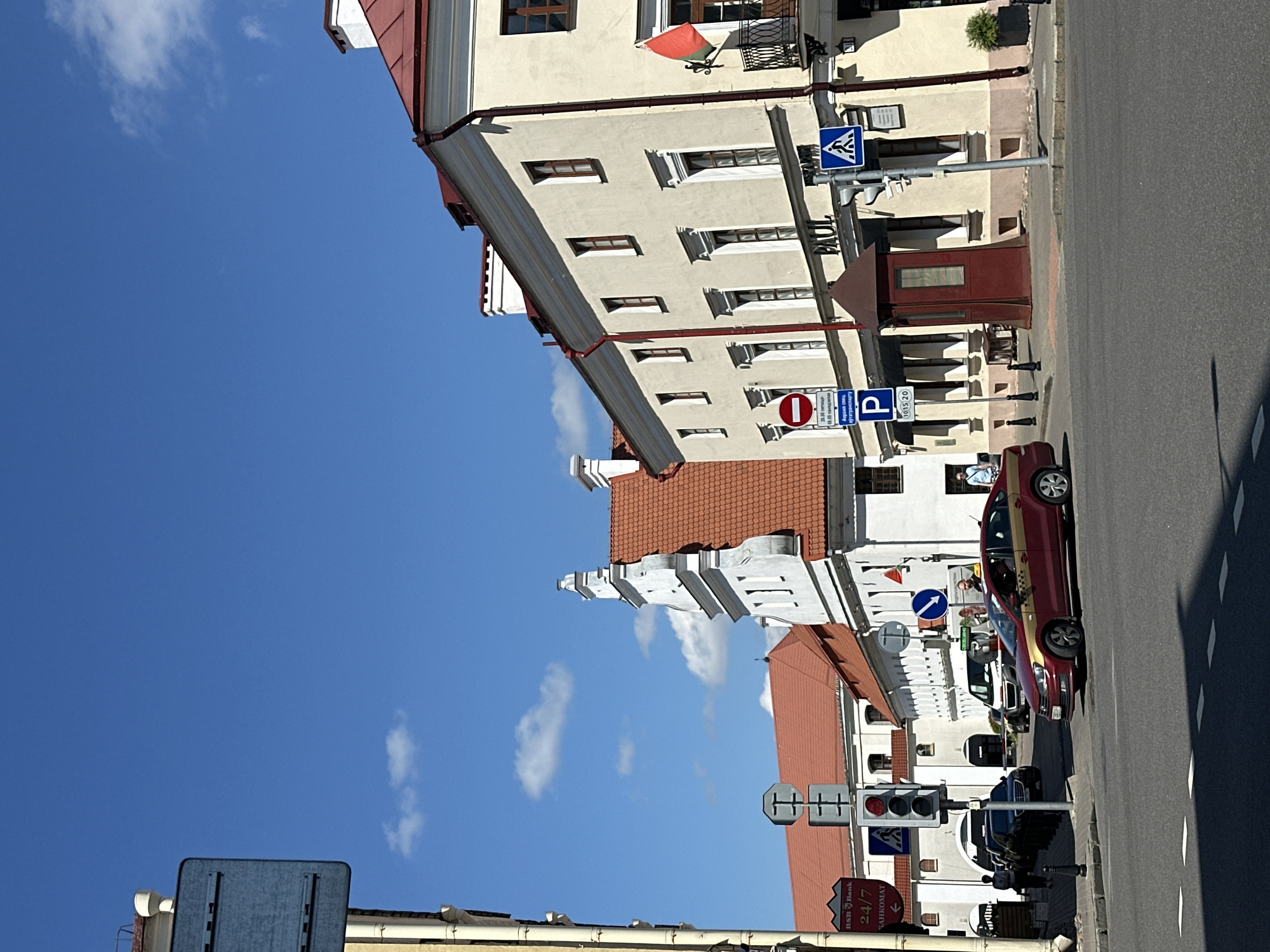 The image captures a street scene in Minsk, Belarus, on a clear, sunny day. In the foreground, a red and gold taxi is turning right at an intersection. Behind the taxi, several other vehicles are stopped at a traffic light. The buildings lining the street feature traditional European architecture with red-tiled roofs and light-colored facades. One building has a prominent sign for "PAUL." Traffic signs, including pedestrian crossings and a parking sign, are visible. A small kiosk with a dark red roof stands on the sidewalk. Several pedestrians are seen walking on the sidewalks and near the buildings. The sky is a vibrant blue with scattered white clouds.