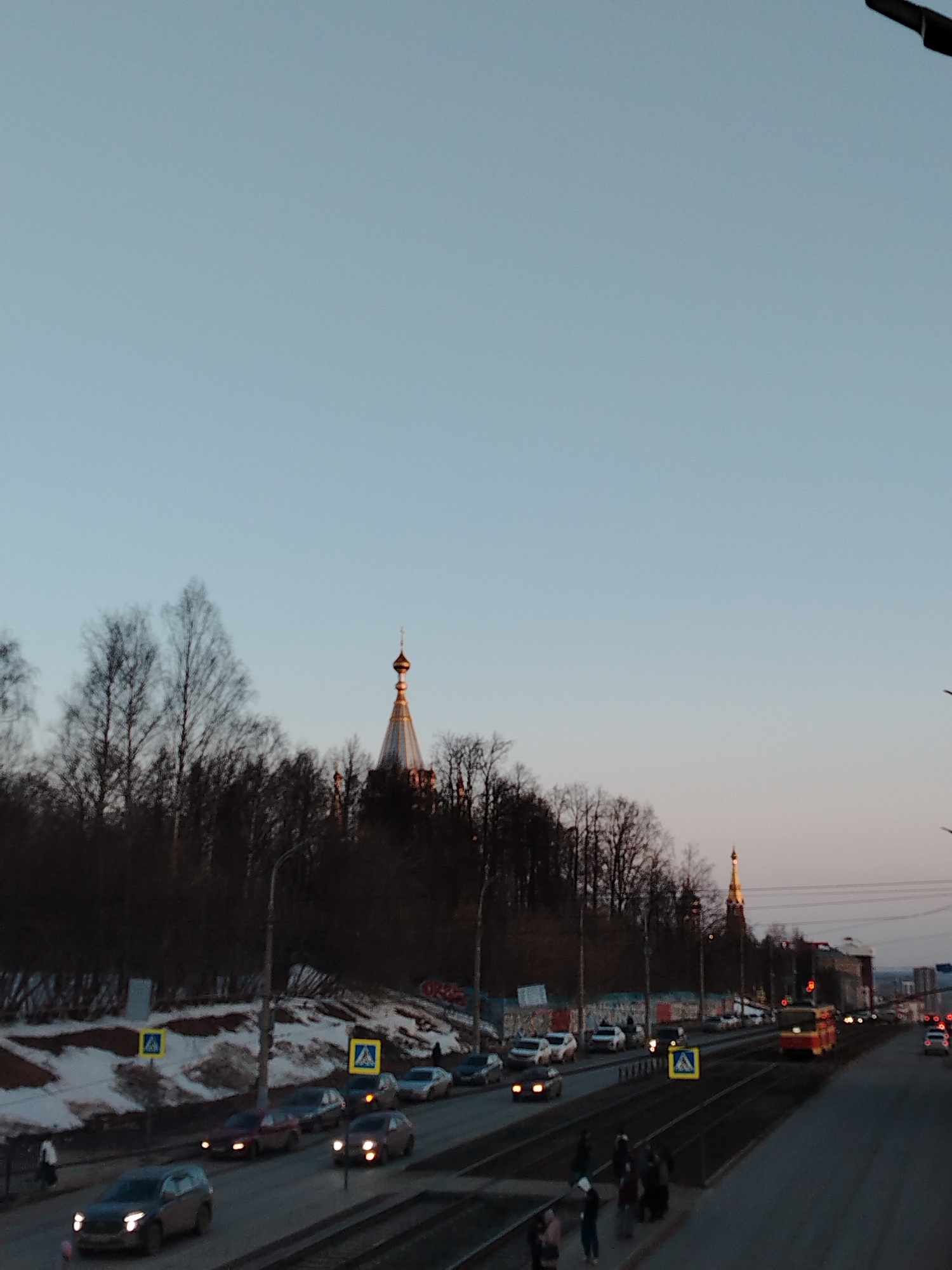 The image captures an urban street scene in Izhevsk, Russia, at twilight under a clear, pale blue sky with a subtle orange glow near the horizon. In the mid-ground, the Holy Dormition Cathedral, identifiable by its prominent golden onion dome and conical spire, rises above a densely vegetated embankment to the left. Patches of residual snow are visible on the ground beneath the bare deciduous trees covering the embankment.
A multi-lane road with active vehicle traffic extends horizontally across the lower and central portions of the frame. Several cars are present on the road, with illuminated headlights indicating low light conditions. Parallel tram tracks run to the right of the main road, accommodating a red and yellow tram traveling away from the viewer. Multiple yellow square pedestrian crossing signs are spaced along the roadside.
In the immediate foreground, several pedestrians are standing on a paved area adjacent to the tram tracks, suggesting a tram stop or designated crossing point. A lone pedestrian walks on a sidewalk further to the left. In the distant background on the right, another smaller golden spire is visible amidst the city skyline.