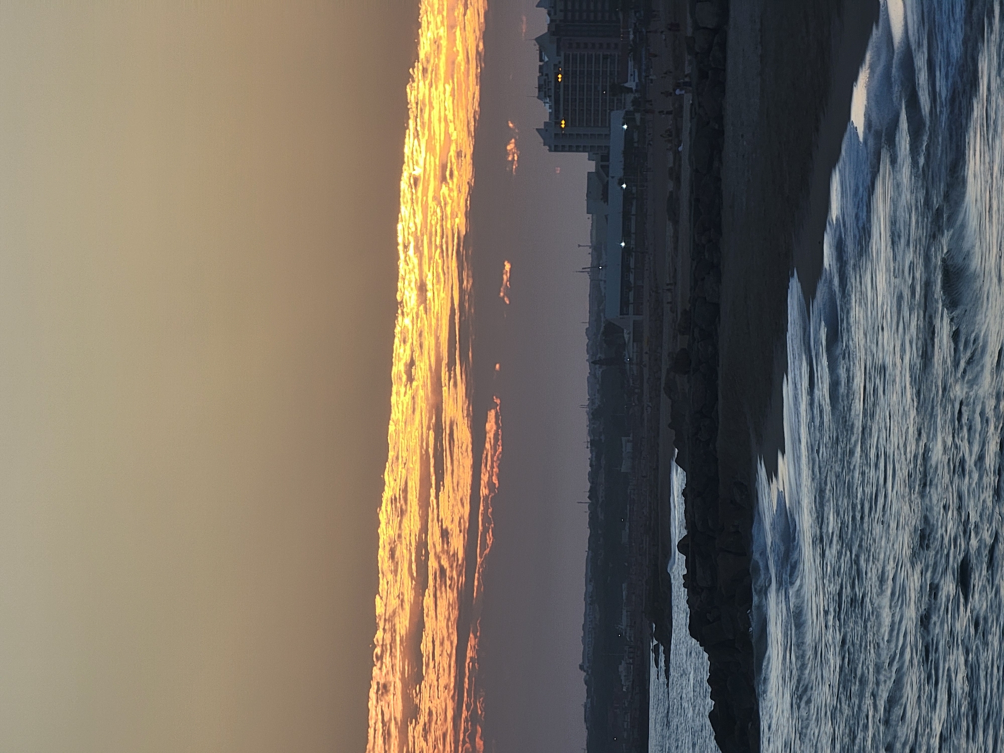 An image captures a sunset over the Atlantic Ocean at Quarteira, Portugal. The sky is a gradient of warm orange and yellow from the setting sun, illuminating a band of textured clouds. Below the sky, a hazy silhouette of the Quarteira coastline is visible, featuring buildings, including a prominent hotel with illuminated windows, and distant infrastructure. In the foreground, dark, angular rocks form a breakwater along the beach. The ocean's surface is a deep blue, with white-capped waves gently rolling towards the sandy shore. A few scattered people are visible on the beach and along the promenade in the middle ground.