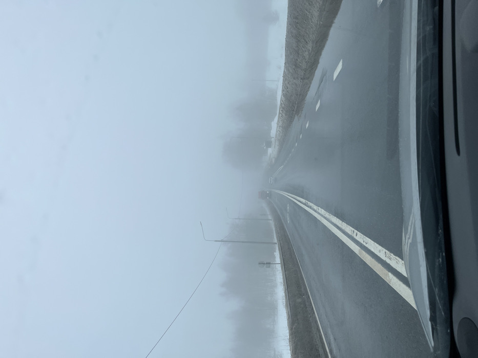 The image displays a two-lane asphalt road in Isakovo, Russia, viewed from inside a vehicle's dashboard perspective. Heavy fog and an overcast sky obscure the distant horizon. The road surface is wet, reflecting ambient light, with a double solid white line separating the left lane and dashed white lines marking the right lane. Snow and dirt accumulation lines both road shoulders, with puddles evident along the right side. Multiple utility poles with streetlights and overhead power lines are visible along the left side of the road, receding into the fog. Bare trees are discernible through the dense fog on both sides of the roadway. In the distance, an oncoming red heavy vehicle, likely a truck or bus, is partially visible, followed by a smaller white vehicle, both significantly obscured by the fog. No individuals or explicit actions are present in the scene.
