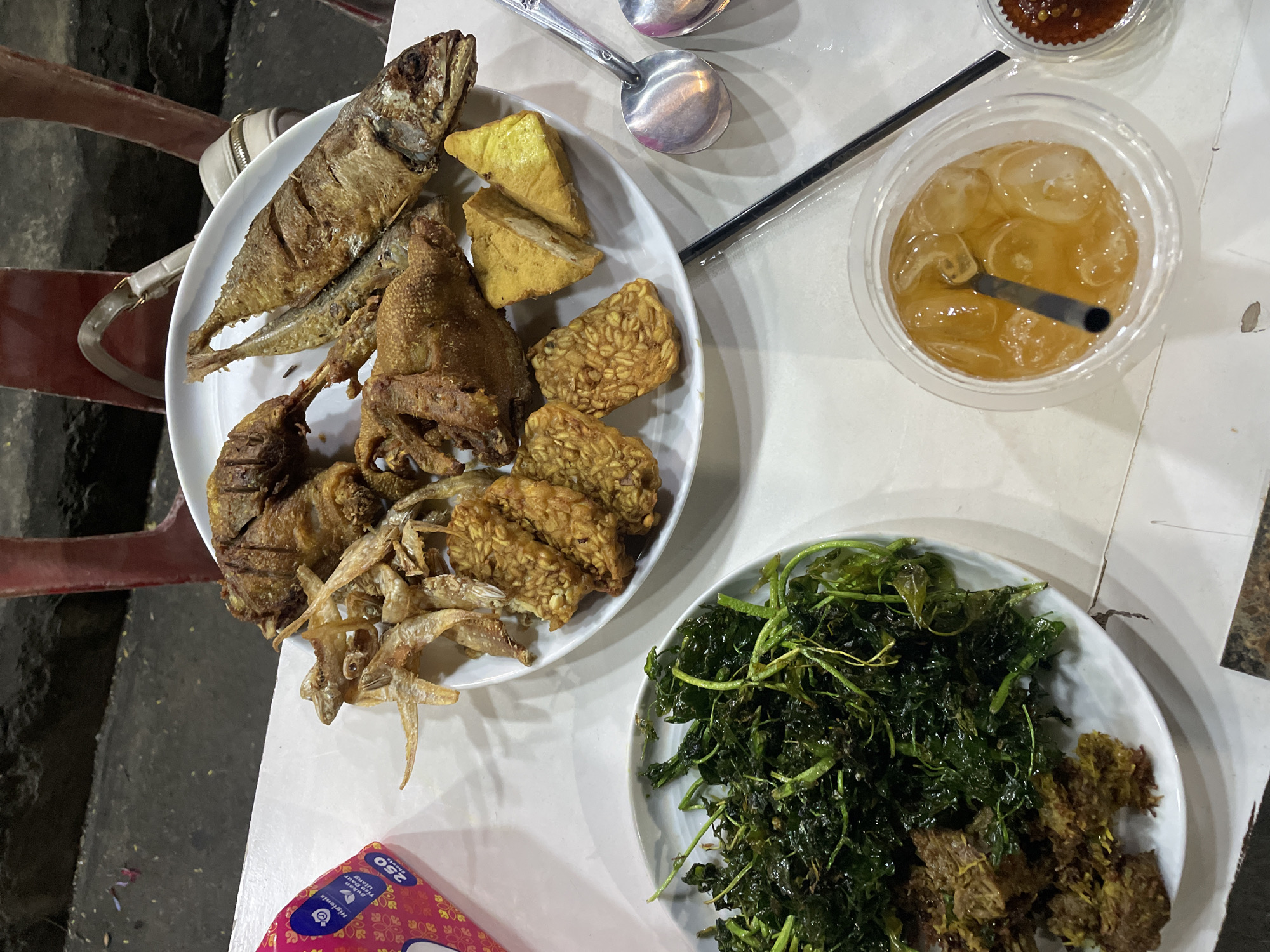 The image shows a meal on a white table, likely in an outdoor or semi-outdoor setting. The meal consists of two white plates. The first plate contains fried fish, fried chicken, fried tofu (tempeh), and small fried anchovies. Silver spoons rest on the table next to the plate. A clear plastic cup containing a light-colored beverage with ice and a black straw sits to the right. To the upper right of the cup is a small container with a red sauce. The second plate has a dark green leafy vegetable dish. There is also another dish with what looks to be some fried food. The background is dark, suggesting it is evening or night. A red chair can be partially seen to the left. A red tissue packet is at the bottom left corner of the frame.
Based on the food items visible, particularly the tempeh and the style of fried dishes, this meal is most likely of Indonesian origin, specifically from Jakarta. It could be sold in one of the many street vendors or restaurants that the area is known for.