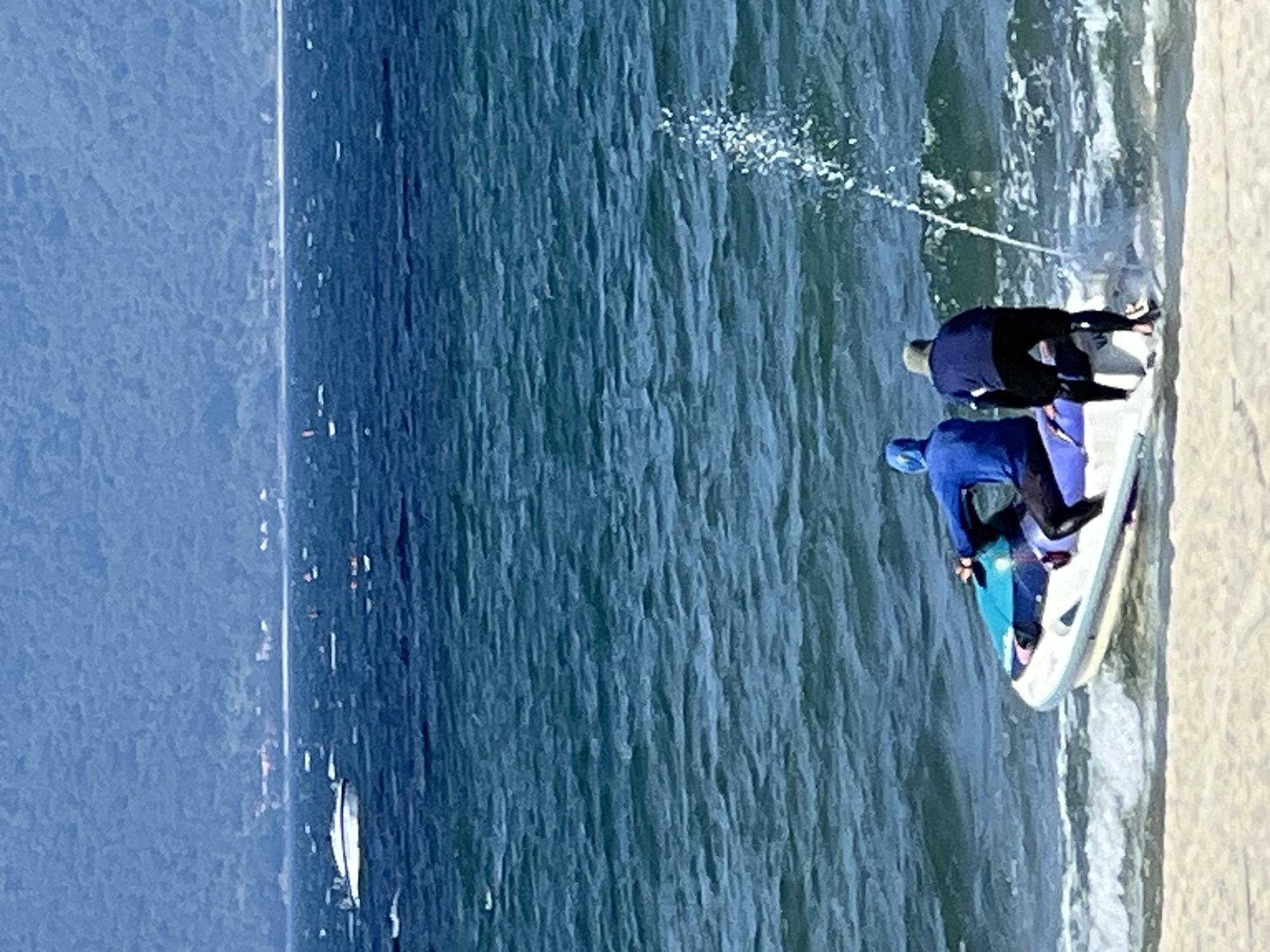 Two individuals are on a jet ski partially on the sandy shore of a beach, with the ocean stretching out before them. The jet ski is white with blue and purple accents. One person, wearing a bright blue hooded jacket and black pants, is seated on the jet ski. The other person, dressed in a dark blue long-sleeved shirt, dark pants, and a light-colored cap, is standing behind the seated individual, facing the water. The jet ski is angled towards the ocean, and a stream of water is spraying from its rear, indicating it is either in operation or has just been. In the background, the deep blue ocean exhibits moderate waves, and distant boats are visible on the water. Beyond the ocean, a hazy, blue-green mountainous landscape with scattered buildings can be seen. The sky is clear and bright. This scene is consistent with the coastal areas of Đà Nẵng, Vietnam, a city known for its extensive beaches and proximity to mountains.