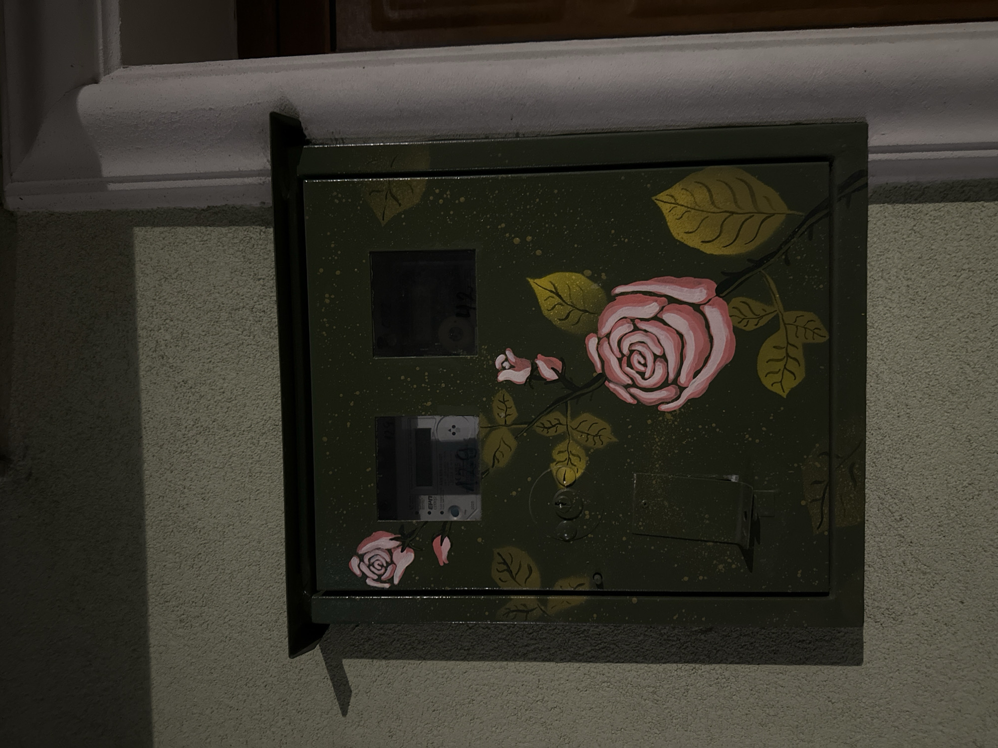 The image shows a green metal electrical box mounted on a textured, light green wall. The box is decorated with painted roses and leaves in shades of pink and yellow. Two small rectangular windows display electrical meters. The scene is likely in Veliko Tarnovo, Bulgaria, as indicated in the prompt, although specific contextual details within the image do not confirm the location. The box is attached below a white ledge. The lighting is dim, creating shadows on the wall and box.
