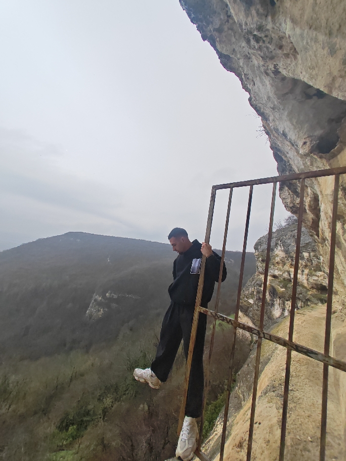 The scene depicts a male individual positioned precariously on a heavily rusted, dark brown metal grate or gate attached to a light-colored limestone cliff face. This location is identified as the Shumen Plateau, near the city of Shumen, Bulgaria, overlooking a deeply forested valley. The male, appearing to be in his late teens or twenties, is dressed in a black hoodie with a light rectangular patch on the left chest, black trousers, and light-colored athletic shoes. He is gripping an upper horizontal bar of the metal structure with his left hand for stability, while his feet rest on a lower horizontal bar. His head is tilted downwards, his gaze directed towards the dense, leafless deciduous forest filling the valley below. The sky is overcast and uniformly grey. The immediate right foreground shows the textured, weathered rock of the cliff face extending upwards and into the frame.