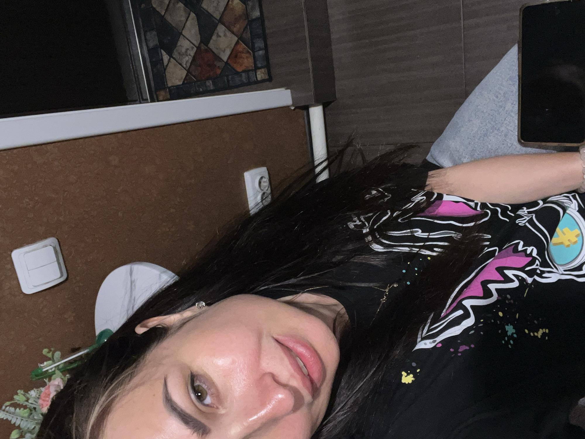 In this close-up selfie, a woman with long, dark hair and a black t-shirt with a graphic print smiles at the camera. She has visible makeup, including blush that highlights her cheeks. Her left eye is prominent, with a light brown iris and visible eyelashes. To her left, a white electrical outlet with two sockets is mounted on a brown, textured wall. A portion of a white frame is visible to the right of the wall, extending towards the top of the frame. Below the frame, a dark surface is present, with a geometric-patterned rug visible in the background. The woman's right arm is partially visible, resting on what appears to be a phone. A white object, possibly a sink, is also partially visible behind her head. The overall lighting suggests an indoor setting.