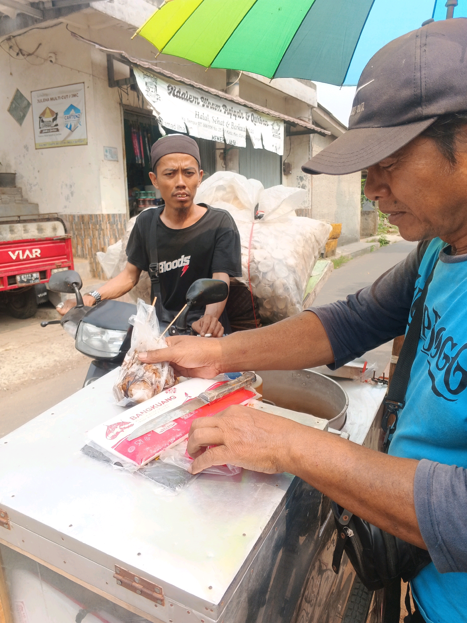 This is a medium shot, eye-level, vertical photograph of two men by a street vendor's cart in Kadu, Indonesia. In the foreground, on the right, a man wearing a blue t-shirt and a black baseball cap is partially visible. His right arm is extended, and his hands are handling some items on the cart. He appears to be preparing or serving food. The cart itself is made of metal and has a hinged door. On the cart, there are packets of food and a knife.
In the midground, to the left, a second man is sitting on a motorcycle and looking at the camera. He is wearing a black t-shirt and a dark cap. He has a bag of food in his right hand. The motorcycle is red and has a license plate. Behind the men, a street scene is visible with buildings and shops. One building has a sign that says "Naalem Viram Rajjo". A colorful umbrella with green and yellow sections is overhead. The ground is paved.