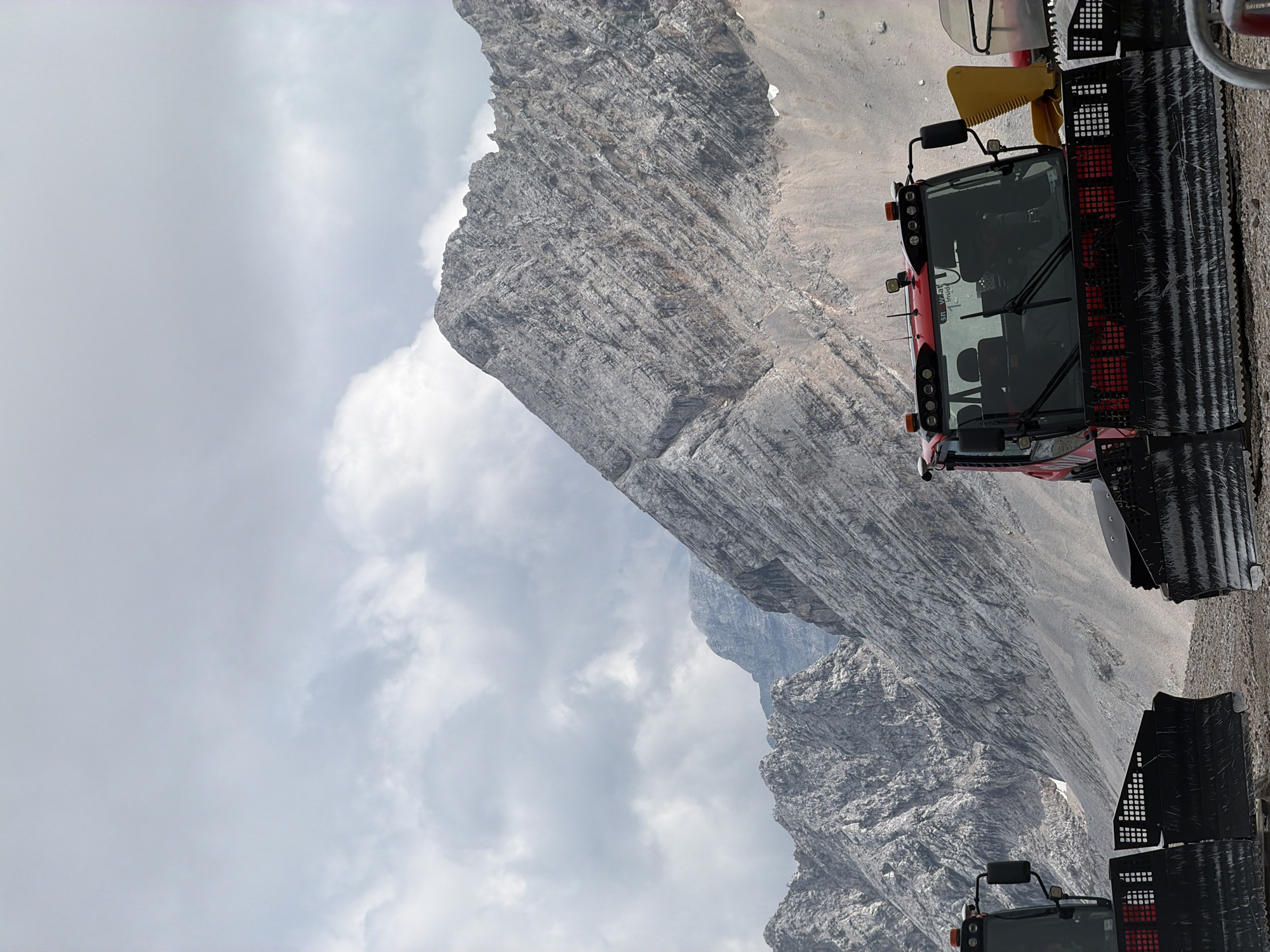 The image depicts a mountainous landscape under a cloudy sky. In the foreground, two red snowcats are partially visible, positioned on a gravelly surface. The snowcats have large black plows at the front. One snowcat on the right is more prominently featured, showing its cabin with windows reflecting the surroundings and various lights on its roof. The background is dominated by rugged, grey mountains with visible striations, suggesting a rocky terrain. The overall scene appears to be an outdoor environment in a high-altitude, possibly alpine, region.
Regarding the location in Garmisch-Partenkirchen, Germany, this scene likely depicts a mountain accessible from the area, such as the Zugspitze or a surrounding peak, where such snow groomers are used for maintaining ski slopes or access routes.