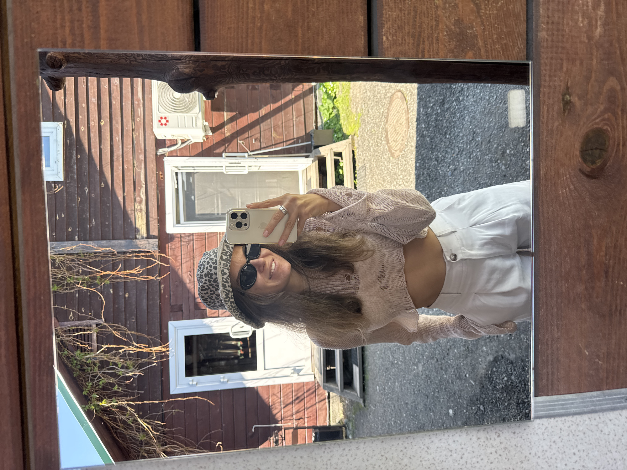 A mirror reflects a young woman taking a selfie with a smartphone. She wears a leopard print bucket hat, sunglasses, a light pink, cropped, holey sweater, and white high-waisted pants. The reflection includes the facade of a two-story wooden building with white-framed windows and an air conditioning unit mounted on the wall. The environment suggests a residential area in Aya, Russia, based on typical building styles and street surfaces found in that region.