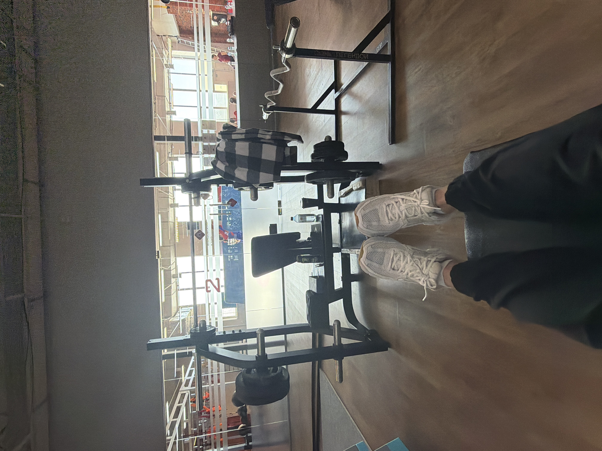 The image captures an indoor gym setting, viewed from a first-person perspective, with the photographer's legs and feet in the foreground. The person is wearing black pants and white athletic sneakers, appearing to be seated on a workout bench or mat.
In the midground, several pieces of black metal gym equipment are visible. This includes a weight rack holding an EZ curl bar and several weight plates. A black and white checkered blanket or towel is draped over one section of the equipment. A clear water bottle rests on the floor nearby.
The background is dominated by large windows that span much of the wall, allowing bright natural light to fill the space. Through these windows, an exterior view of another building or structure can be seen, with some faint human figures and scaffolding-like elements. The lighting suggests it is daytime.
Visible text on the gym equipment in Cyrillic reads "СПОРТ ЛЕГЕНДОЙ" (SPORT LEGEND) and "СЛР" (possibly an abbreviation like Sports League of Russia). Faintly visible through the windows, on what appears to be a sign or poster, is partially obscured Cyrillic text including "ВОРОНЕЖСКОЙ КАМБ" and a prominent red character resembling an 'N' or 'И'.
The overall scene depicts a moment of rest or preparation within a modern, well-lit fitness center equipped for weight training, located in Chaikovsky, Russia.