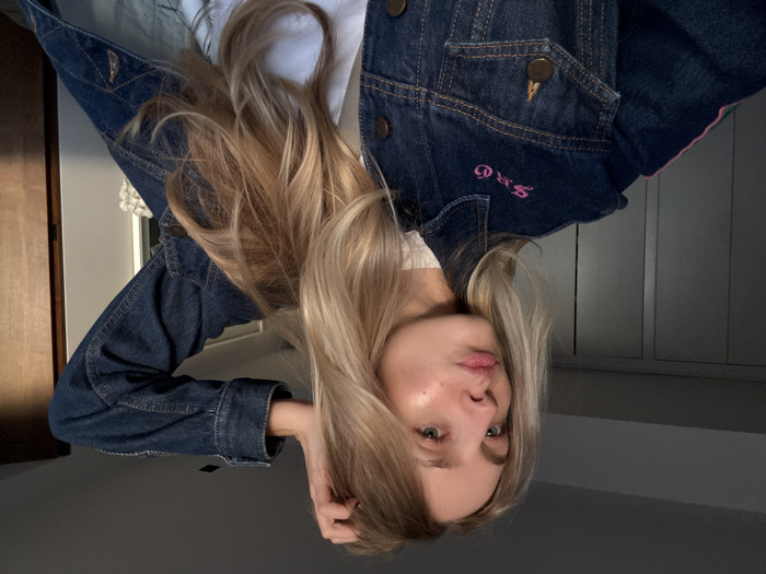 A young adult female subject with long, wavy blonde hair and light eyes is shown in an indoor setting. Her lips are pursed into a pout, and her right hand is raised, with fingers lightly touching her hair near her temple. She is wearing a blue denim jacket over a white top. The denim jacket features "SRG" embroidered in pink script on the upper left chest area and a vertical pink stripe along the upper left sleeve. The background consists of a plain wall, appearing to be a light gray or off-white, with vertical seams suggesting a closet or wardrobe on the left. A darker, possibly wooden, vertical surface is visible on the far right. Warm, natural light illuminates the subject from the left side, highlighting her face and hair. The specific location within Mexico City, Mexico, cannot be determined from the image content.