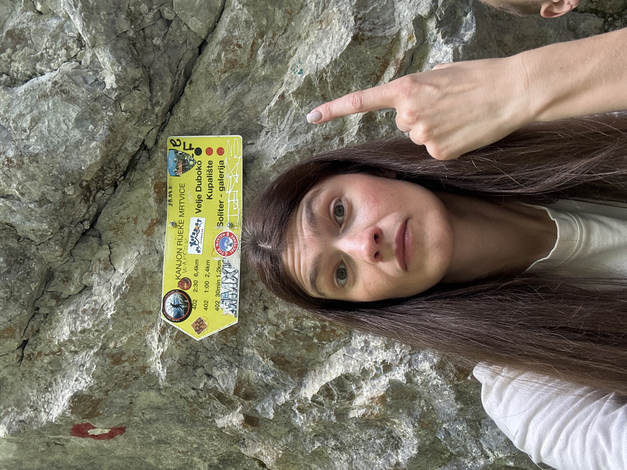 A woman with long brown hair points toward a sign affixed to a textured gray rock face. The sign, primarily yellow, features information about hiking trails, including distances and estimated times. Text on the sign indicates locations such as "KANJON RIJEKE MRTVICE," "Velje Duboko," "Kupalište," and "Soliter - galerija." A red and white trail marker is visible on the rock face beneath the sign. A hand partially obscures the top right corner of the frame. The image is rotated, presenting a sideways view of the scene. The image appears to have been taken in Lazba, Montenegro.