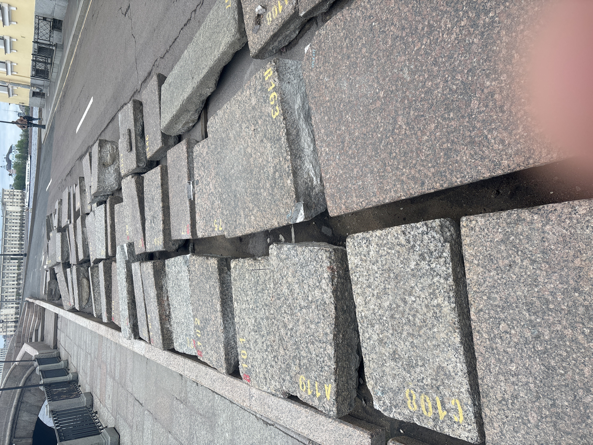 The image shows a row of granite blocks with yellow lettering placed along a sidewalk near a road in Saint Petersburg, Russia. The blocks are laid out next to a lower walkway with a metal railing, potentially alongside a canal or river based on the presence of water and distant buildings visible in the background. Two people walk down the road in the distance. The photograph is taken at an angle, causing a distorted perspective. The environment suggests a historical and architectural context typical of Saint Petersburg.