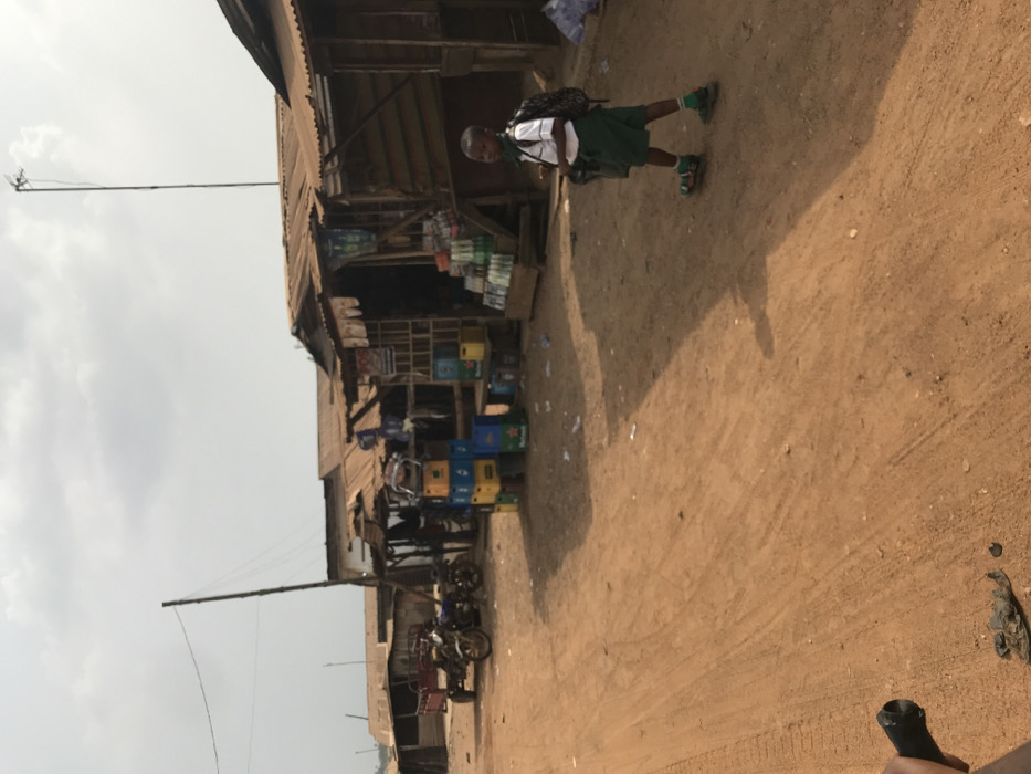 The scene depicts a rural street in Ogbese, Nigeria, under an overcast sky. A young male child, dressed in a white short-sleeved shirt, green shorts, green socks, and sandals, walks right-to-left across a reddish-brown dirt road. He wears a dark backpack and carries a green bag in his left hand. To the child's left, a row of market stalls constructed from wood planks with corrugated metal roofs lines the street. In front of these stalls, stacks of plastic crates, some bearing "Heineken" branding in blue, yellow, and green, are present. A display stand holds numerous bottled beverages. Further left, two motorcycles are parked next to a small, red two-wheeled trailer. A tall, slender pole with wires extends from the left side of the frame, and a similar antenna-like structure is visible further back on the right. Scattered debris and litter are visible on the ground. A dark object, possibly part of a bicycle or a hand, is partially visible in the bottom left foreground, suggesting the photographer's perspective.