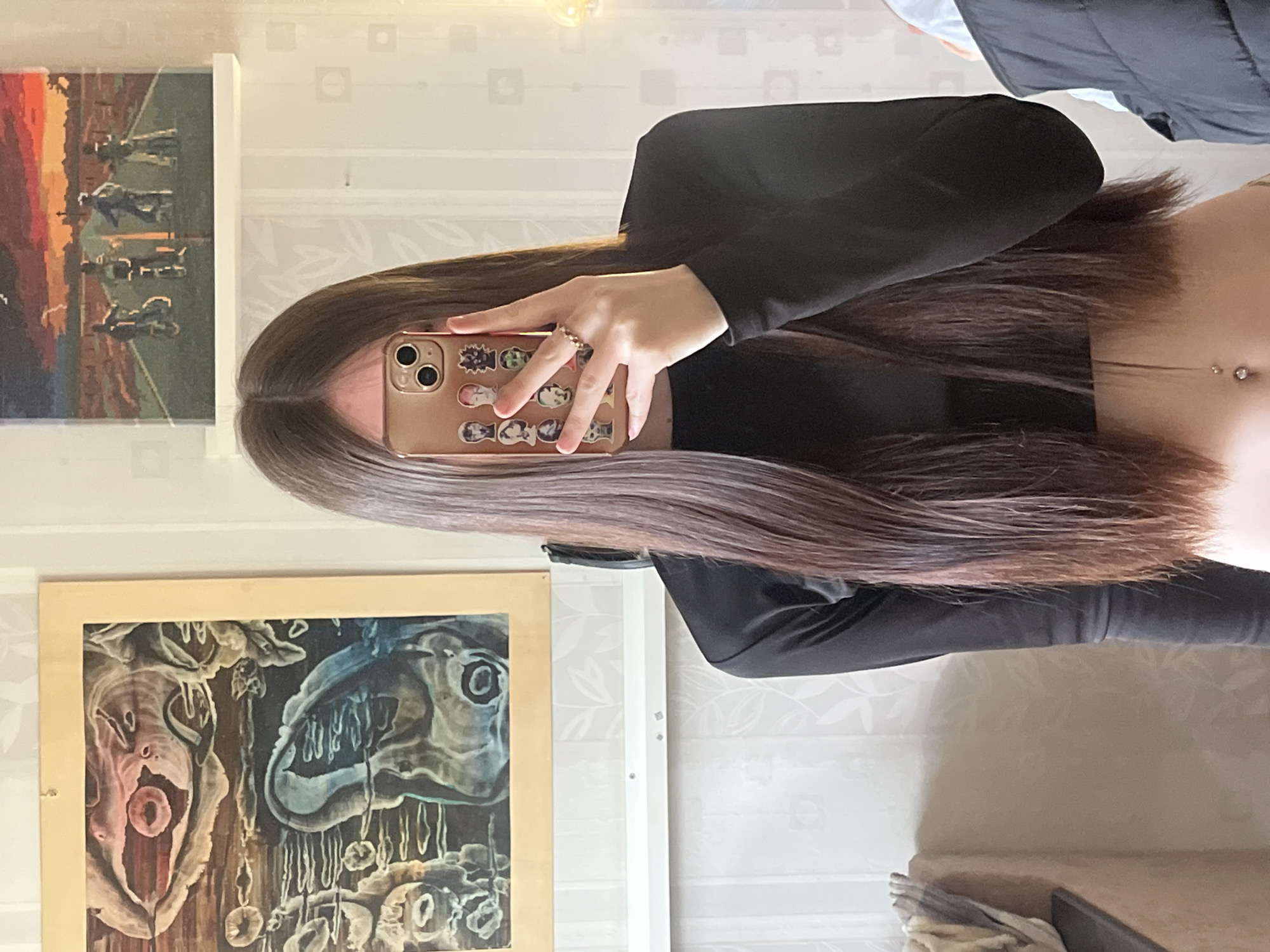 A woman with long brown hair holds a phone with a tan case decorated with cartoon stickers in front of her face, partially obscuring it. She wears a black top. A piece of jewelry is visible on her stomach. Two paintings hang on the wall behind her. The background consists of a wall with a light-colored, subtle pattern and furniture. The image suggests a domestic setting, likely in a private residence of an unknown location in Russia.
