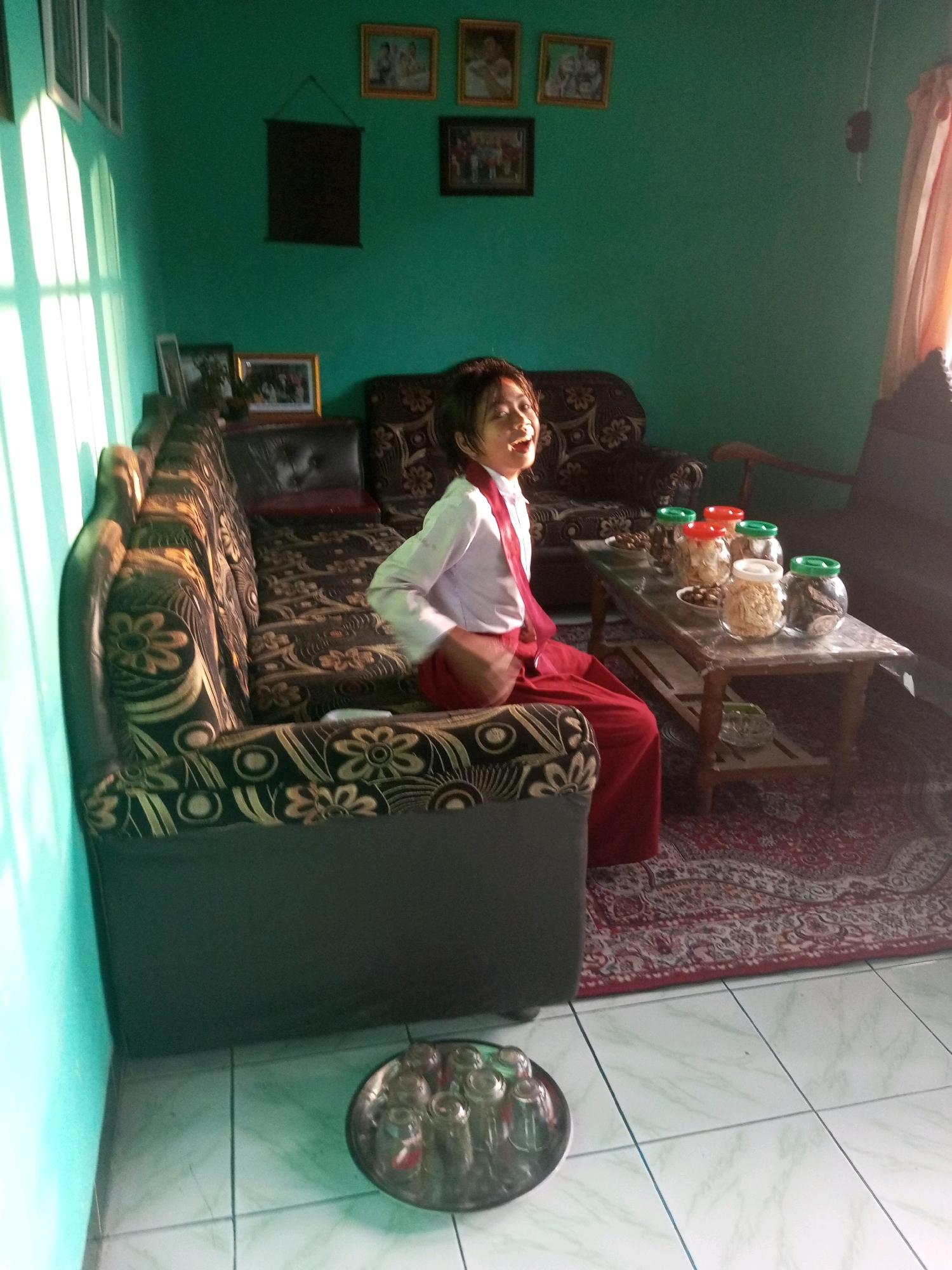 In the city of Karangsari, Indonesia, a young person wearing a white shirt with a red tie and red skirt is sitting on a patterned sofa. The sofa is positioned against a turquoise wall. The young person is looking to their right and smiling. In front of the sofa, a wooden table holds several jars filled with snacks, and a bowl. Sunlight streams in from the left, casting shadows on the wall. To the right of the sofa, another piece of furniture is visible, and a patterned rug covers the floor. A tray of glasses is placed on the tiled floor in the foreground. The room also contains several framed pictures on the wall, adding to the domestic setting.
