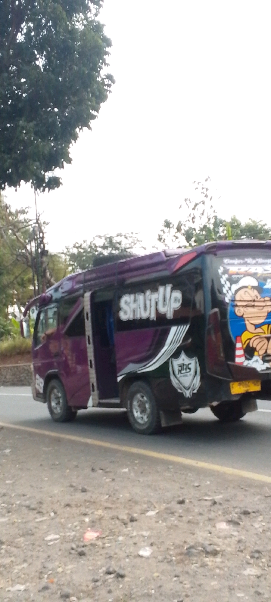 A purple and black van with the words "SHUT UP" printed on its side is parked on the side of a road. The van has its doors open. On the rear of the van, a cartoon character that resembles Popeye is painted, holding a pipe. The van is parked in a rural or suburban setting, with trees and foliage visible in the background. The road appears to be made of asphalt and there is debris on the side of the road. The sky is overcast.