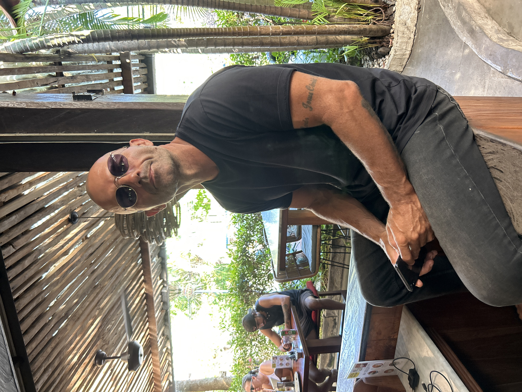 A man with a shaved head and sunglasses is seated outdoors, looking at the camera. He is wearing a black t-shirt and dark jeans. His right arm is visible, showing a tattoo that reads "Jesus Christ". He is holding a black smartphone in his hands. The setting appears to be a cafe or restaurant with a wooden ceiling structure, palm trees, and other patrons at tables in the background. The scene suggests a relaxed, tropical environment, possibly in North Kuta, Indonesia, given the vegetation and open-air architecture.