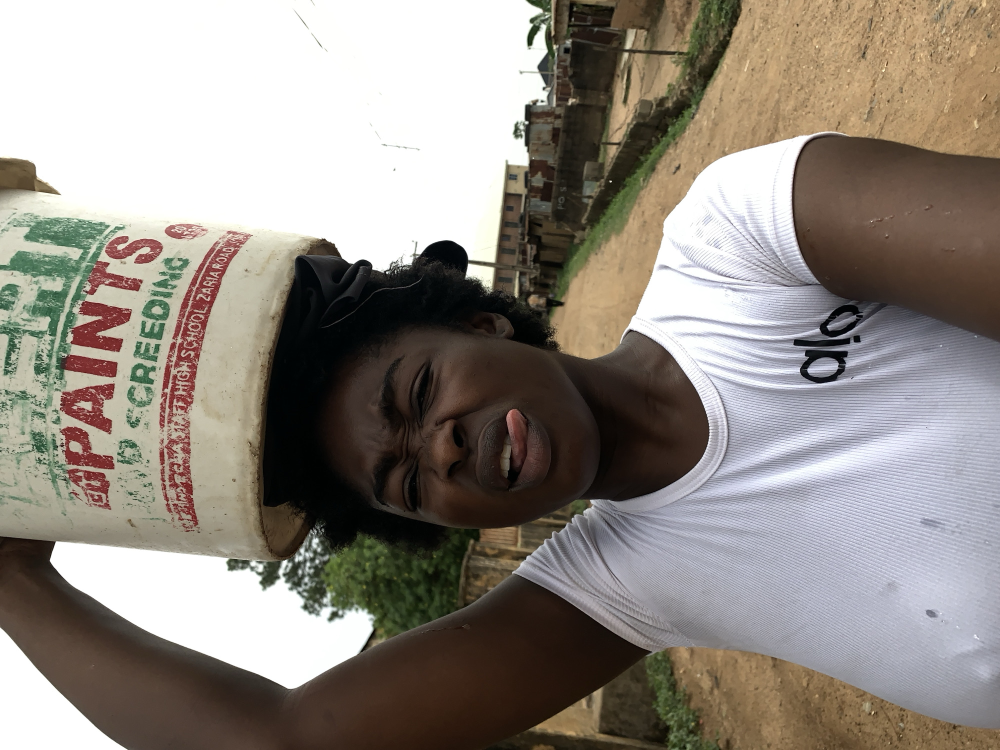 The image shows a woman in Rafin Par, Nigeria, carrying a white bucket labeled "Paints" on her head. The bucket is positioned directly over her head and obscures the top of her head and forehead. She wears a white tank top with the brand "alo" visible on the chest. Her face shows a strained expression with her tongue sticking out. The background includes a dirt road, vegetation, and buildings. The sky is overcast.