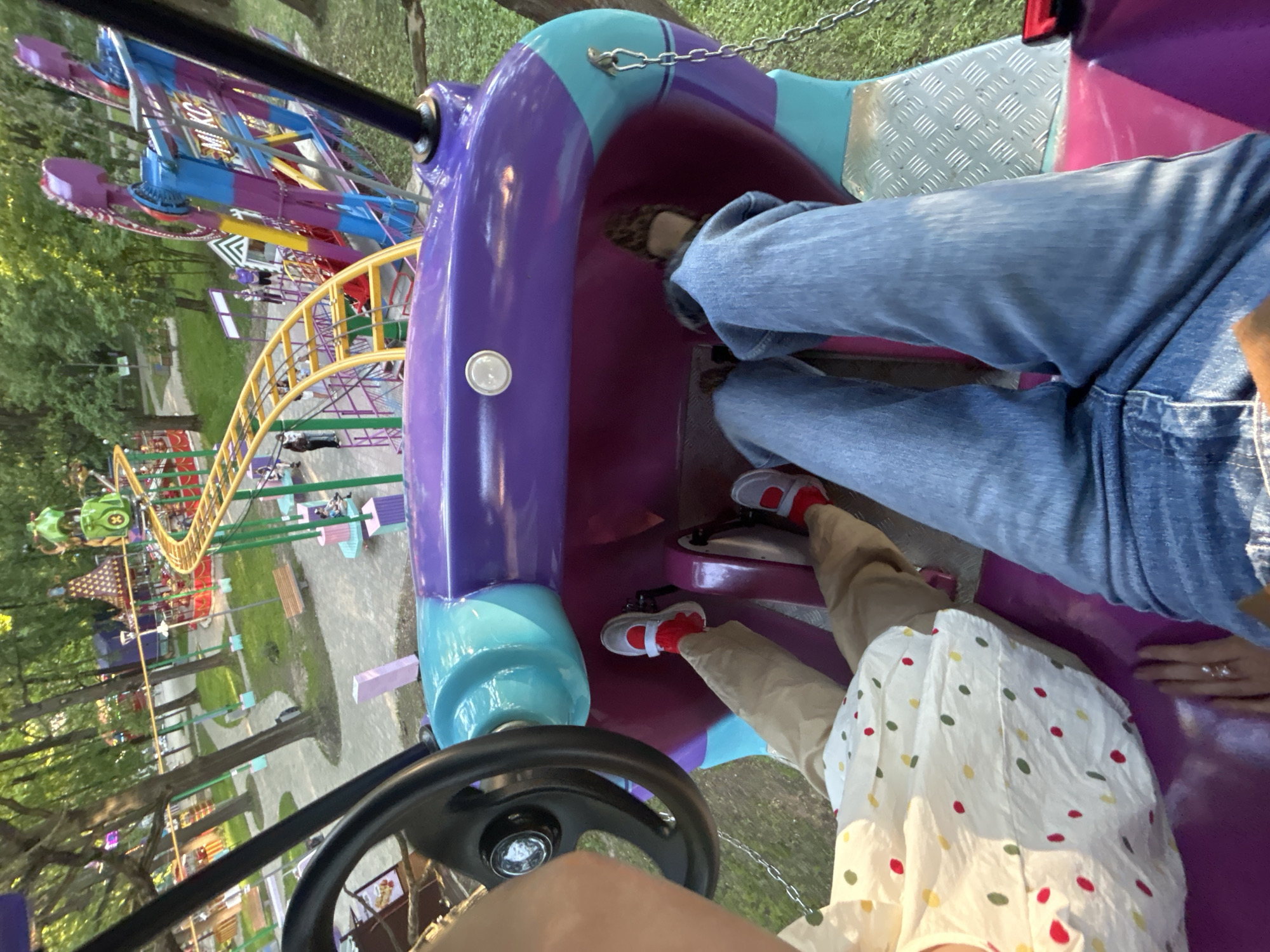 The image provides an elevated, first-person perspective from within a two-person amusement park ride in the city of Khimki, Russia. The ride's interior is predominantly purple with teal blue accents, featuring a black steering wheel on the left and pedal mechanisms for both occupants. Chains hang from the ride's exterior structure on the right.
In the immediate foreground, two individuals are seated. On the left, a person is wearing a white top with red, yellow, and green polka dots, light brown pants, red socks, and light grey shoes with a white sole and a velcro strap. Their left hand rests on the steering wheel. On the right, a second person is wearing blue denim jeans, red socks, and similar light grey shoes. Their right hand, adorned with a ring featuring three small, light-colored stones (possibly pearls), rests on the purple surface of the ride. Both individuals' feet are positioned above the pedal areas. A metallic, diamond-plate textured footrest is visible on the right floor panel of the ride.
Beyond the foreground ride, the amusement park extends into the mid-ground and background. A prominent yellow-tracked roller coaster with green support pillars zigzags through the left-center of the scene, featuring a small green vehicle resembling a frog or bug on its track. Adjacent to this, in the mid-ground right, stands a large, multi-colored swing ride with substantial blue, purple, and yellow components. The partial text "MI KC" is visible on one of its structural arms.
The park environment is characterized by extensive green grass, numerous mature trees with dense green foliage, and paved pathways where several other park visitors are observed walking or standing. Benches are situated along the pathways. Various other smaller, colorful amusement structures and tent-like attractions are visible in the deeper background, indicating a diverse array of park offerings. The scene is brightly lit by natural daylight.