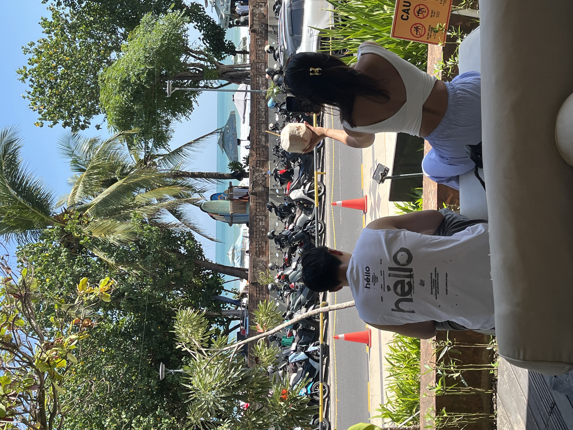 In Kuta, Indonesia, two individuals are seated at an outdoor cafe with a street and beach view. The person in the foreground, facing away from the camera, wears a white tank top with the word "héllo" printed on it, and gray pants. To their right, another person, also facing away, wears a white crop top and light blue striped pants. They are holding a coconut with a straw and are in the process of taking a photo of it with a phone. In the background, a row of parked scooters and motorcycles lines the street. Beyond the street, the turquoise ocean is visible with a few umbrellas and surfboards on the sandy beach. Lush green trees and palm trees provide shade and frame the scene. The sky is clear and blue, indicating a sunny day. A caution sign with pictograms is visible on the right side of the image.