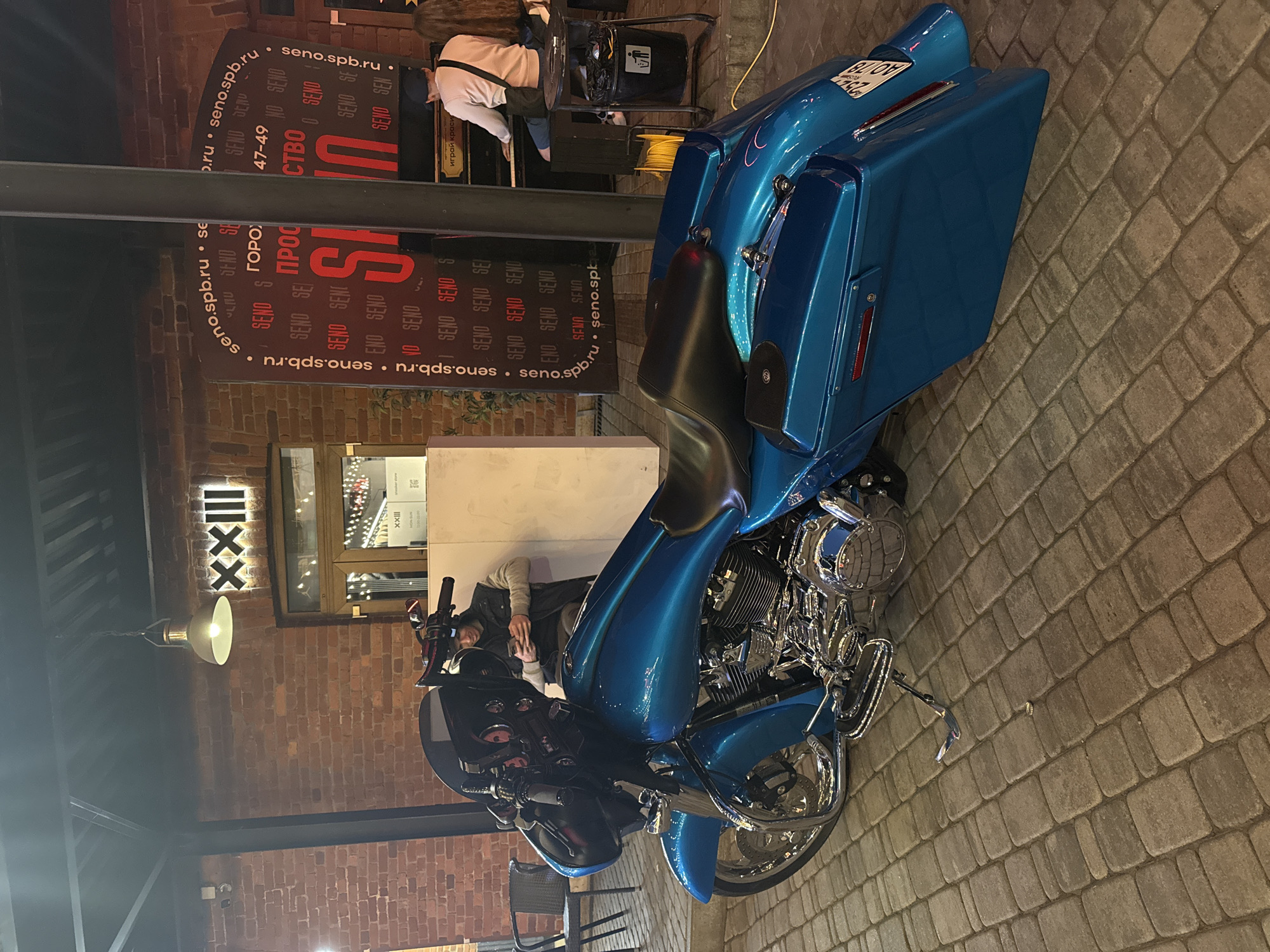 The image depicts a turquoise custom motorcycle parked on a brick-paved outdoor area in front of a building. The building's facade features exposed brickwork and a sign that reads "XXIII". To the right, a large black banner with red lettering displays "SENOP.SPB.RU" repeatedly, interspersed with other text. A man in a dark vest over a gray hoodie is standing in front of a counter, looking at his phone, and a person with long hair is seated at a piano, playing. The lighting suggests it is evening or night. The setting appears to be an urban environment, likely St. Petersburg, Russia, given the ".SPB.RU" domain on the banner. The motorcycle is the primary subject, detailed with chrome accents and custom saddlebags. The ground is paved with interlocking rectangular bricks.