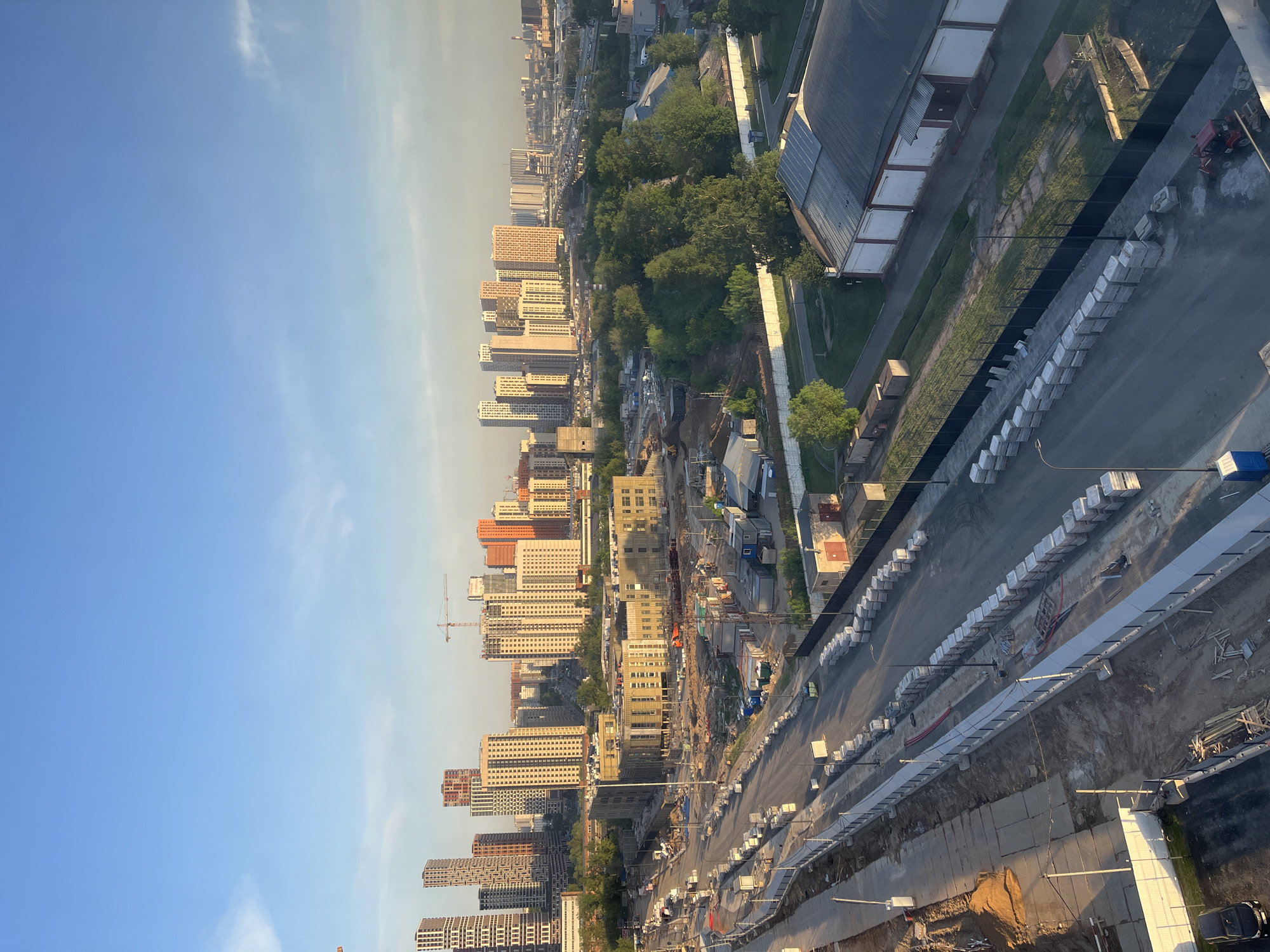 The image depicts an urban landscape, likely Moscow, Russia, viewed from an elevated position. The scene includes numerous high-rise residential buildings of varying colors, primarily white, orange, and beige, stretching along the skyline. A construction site is visible in the foreground, featuring earthworks, construction equipment, and temporary structures. A row of white concrete blocks lines one side of the construction zone, possibly acting as a barrier or temporary sidewalk. Adjacent to the construction site, a roadway contains vehicular traffic. Beyond the buildings, patches of greenery and trees are visible. The sky is clear blue, indicating daytime. In the upper right portion of the frame, a building with a metallic roof is partially visible, possibly part of an industrial or commercial complex.