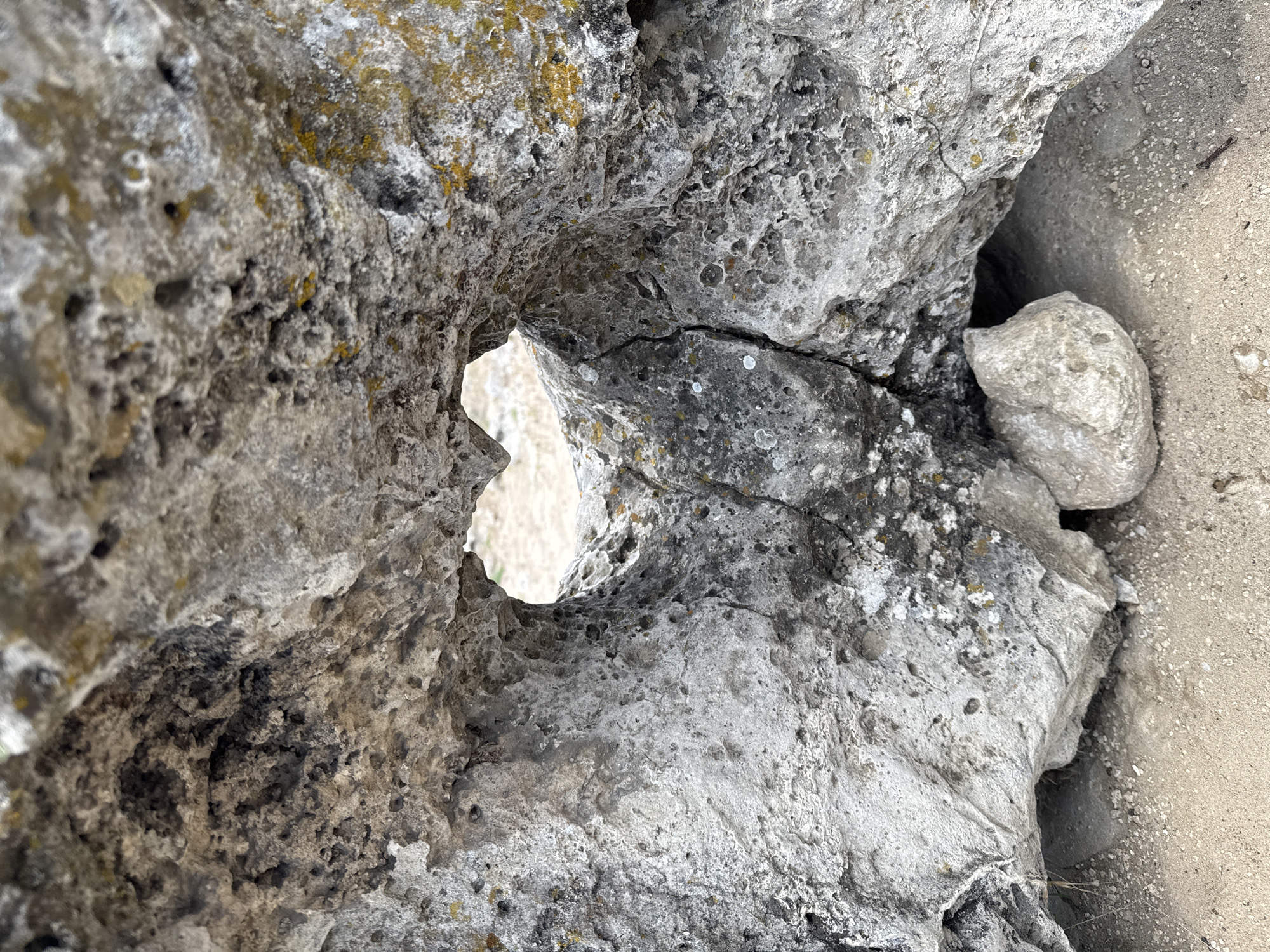A close-up, high-angle shot captures a natural rock formation featuring a distinct, irregularly shaped opening. The dominant subject is a light grey rock with a highly textured, porous, and weathered surface. Patches of dark grey, black, and yellow-green lichen are visible across its surface, suggesting exposure to the elements over time. Several cracks spiderweb through the rock, notably near the central opening.
The opening, positioned slightly to the left of the image center, resembles an abstract heart or keyhole shape, providing a glimpse of a lighter, out-of-focus background, likely sand or dry earth. To the right, another smooth, light-colored rock or boulder is partially visible, nestled against a patch of sandy, light brown or beige ground that extends towards the top and right edges of the frame. The overall lighting suggests a clear daytime, with soft, ambient illumination that highlights the rock's intricate textures without casting harsh shadows. No human activity, specific events, or visible text are present in the image. The scene conveys a sense of rugged natural beauty and geological detail.