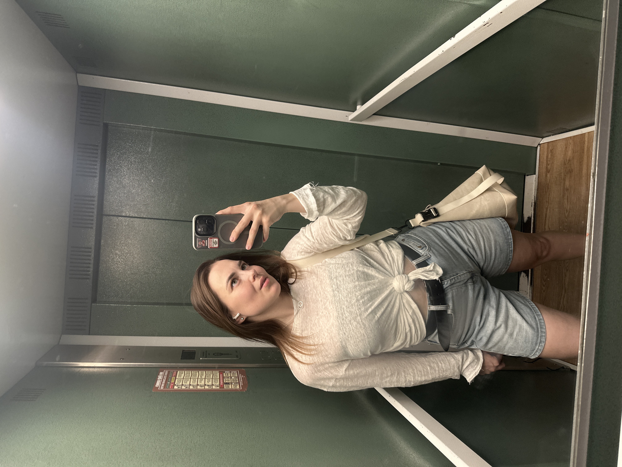 A young woman takes a selfie in an elevator with green walls and a wood-paneled floor. She wears a long-sleeved white shirt tied at the waist, denim shorts with a black belt, and carries a cream-colored canvas bag. The elevator appears to be located in Perm, Russia based on the sign on the wall containing Russian text.