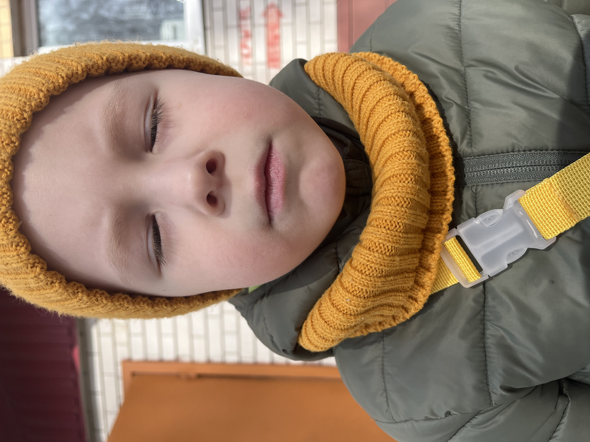A young child with closed eyes is seen wearing a mustard yellow knit hat and matching scarf. The child is also wearing a green quilted jacket with a yellow strap and a white plastic buckle. The background shows an orange-colored object and a wall composed of white and red bricks, suggesting an outdoor setting. The photo was taken in Usman, Russia.