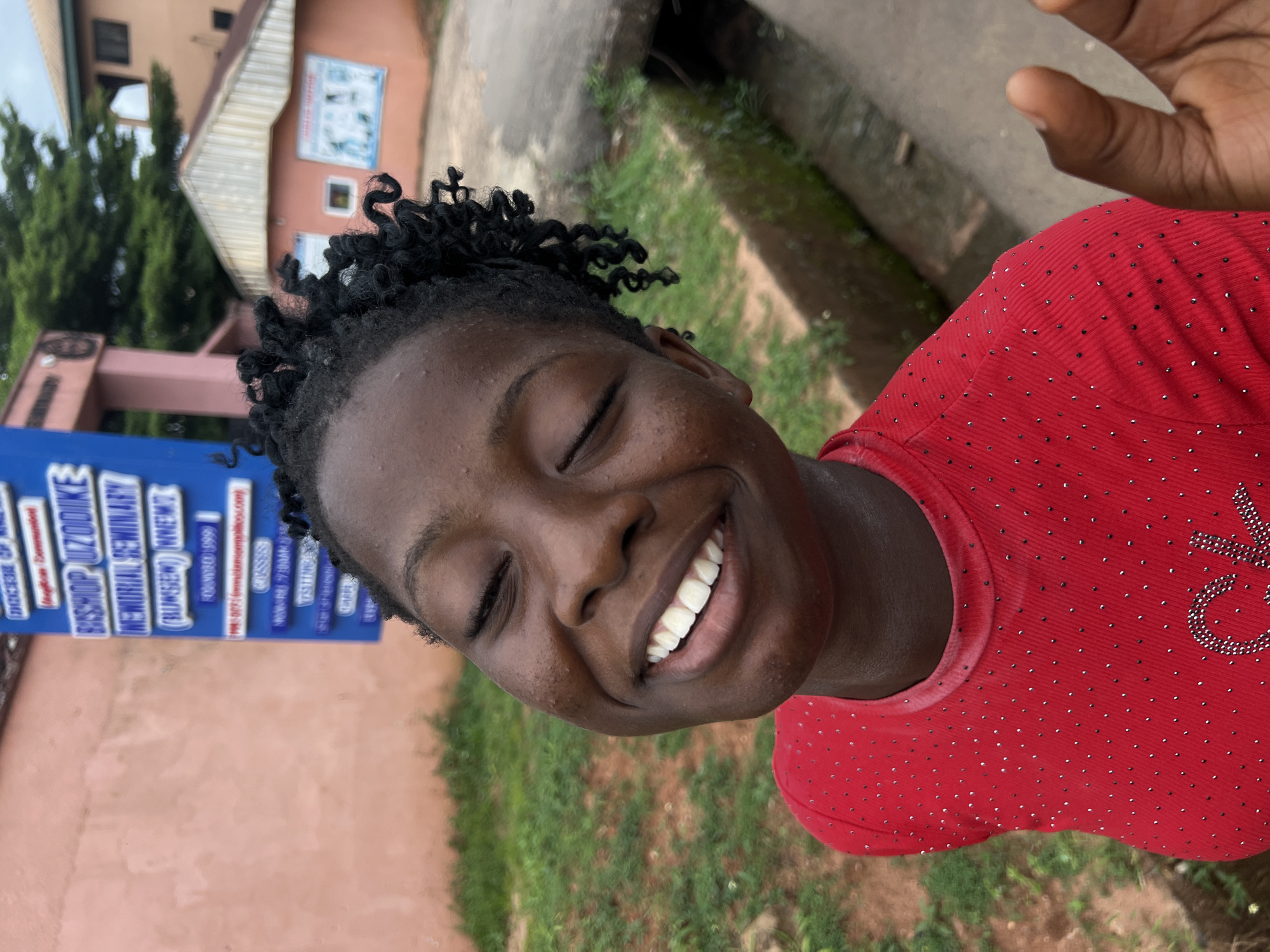 A young Black female with a wide smile and closed eyes, her head tilted slightly upwards, is captured in a medium close-up shot outdoors. She has dark skin and her hair is styled in short, curly dreadlocks. She is wearing a vibrant red shirt adorned with small, shimmering embellishments and a larger sparkling design on the chest. In the background, a blue signpost with white lettering is visible, indicating "Bishop Uzodike Memorial Seminary" and other details, suggesting the location is within the seminary's grounds. To the right, a portion of a light-colored building with a brown roof and a smaller, pink building with a similar roof are discernible. Green grass and a concrete ditch are also part of the environment. The overall scene is one of joy and natural beauty. The signpost confirms the location as Bishop Uzodike Memorial Seminary in Nnewi, Nigeria.