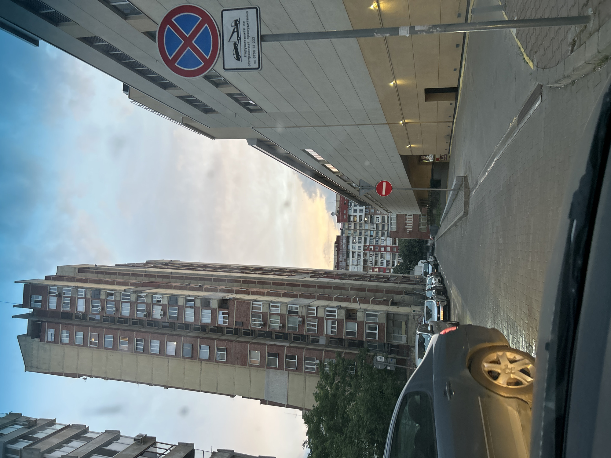 A view from inside a vehicle looking out at a city street. The street is paved with cobblestones. On the left, a tall, narrow, brick-faced apartment building with many windows is visible. Further down the street, another row of apartment buildings can be seen. On the right side of the street, a large, modern building with a beige facade is present. A sign with a red circle and a blue 'X' inside, indicating no stopping, is mounted on a pole in front of this building. Below it, another sign shows a tow truck and text in Bulgarian, with a phone number. A red circular sign with a white horizontal bar is also visible on a pole further down the right side of the street, indicating no entry. Several cars are parked or driving down the street. The sky is partly cloudy with a hint of sunset colors in the distance.