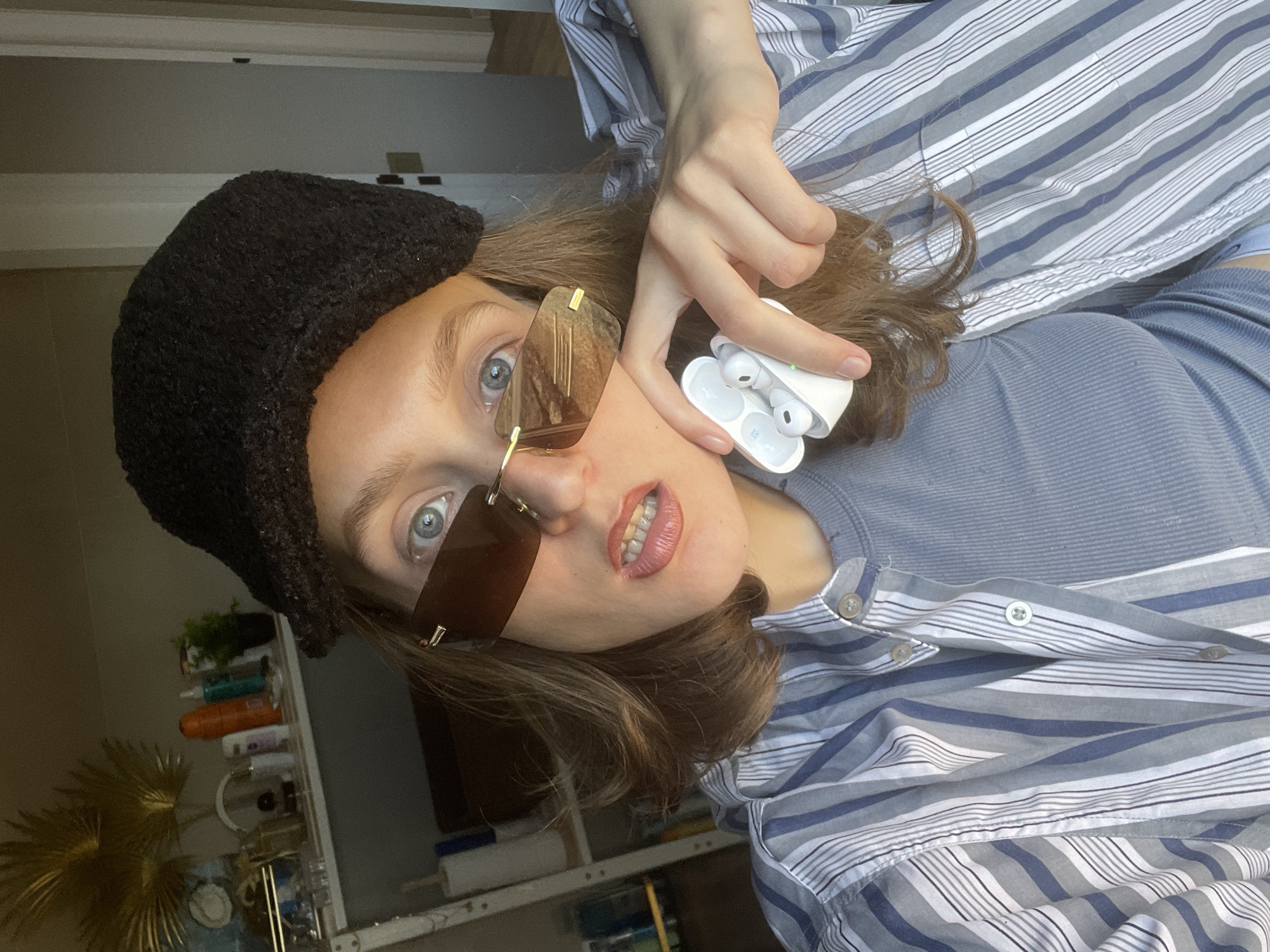 The image is a selfie-style photograph of a young woman. She is wearing a black knit hat, brown-tinted sunglasses with a gold frame, and a striped collared shirt over a blue-gray t-shirt. She is holding a white charging case with two wireless earbuds in her right hand near her face. The background reveals a room with shelves displaying various objects and a decorative gold palm frond. The location is unspecified, but the user provided Sepych, Russia as a possible location.