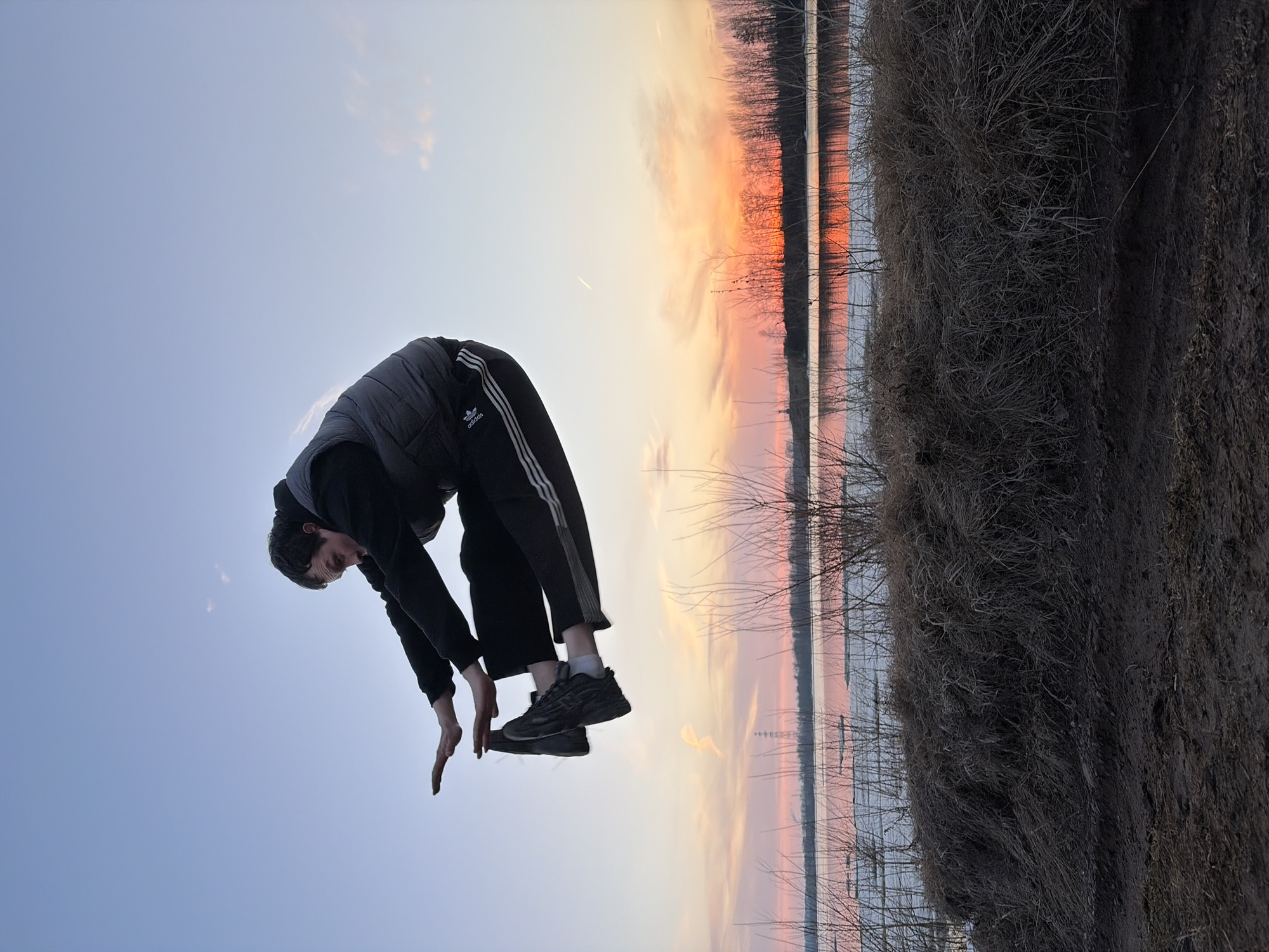 A person is in mid-air, bent over with hands reaching down towards the bottom of the frame. The person is wearing a dark hooded sweatshirt, a gray vest, and black Adidas track pants with white stripes. They are also wearing black athletic shoes. In the background is a sunset reflected in the water, with dark trees lining the horizon. To the right is a mass of dark, tangled vegetation and dirt. The sky is visible in the upper-left corner, ranging from pale blue to orange as it meets the sunset colors. The location is Chaikovsky, Russia.
