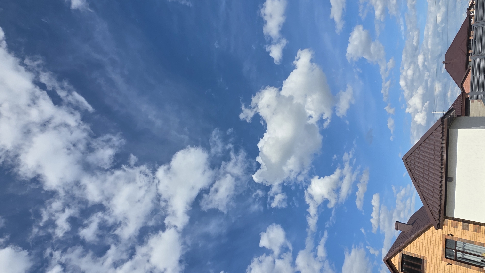 The image shows a view of the sky with clouds, alongside the roof of a building. The sky is a deep blue color with varying cloud formations, including wispy, scattered clouds and denser, puffy white clouds. The building's roof is brown and appears to be made of roofing tiles. The building itself has brickwork visible on the side. There is no evidence to confirm that this photo was taken in the city of Bulgakovo, Russia. There are no people visible in this scene.