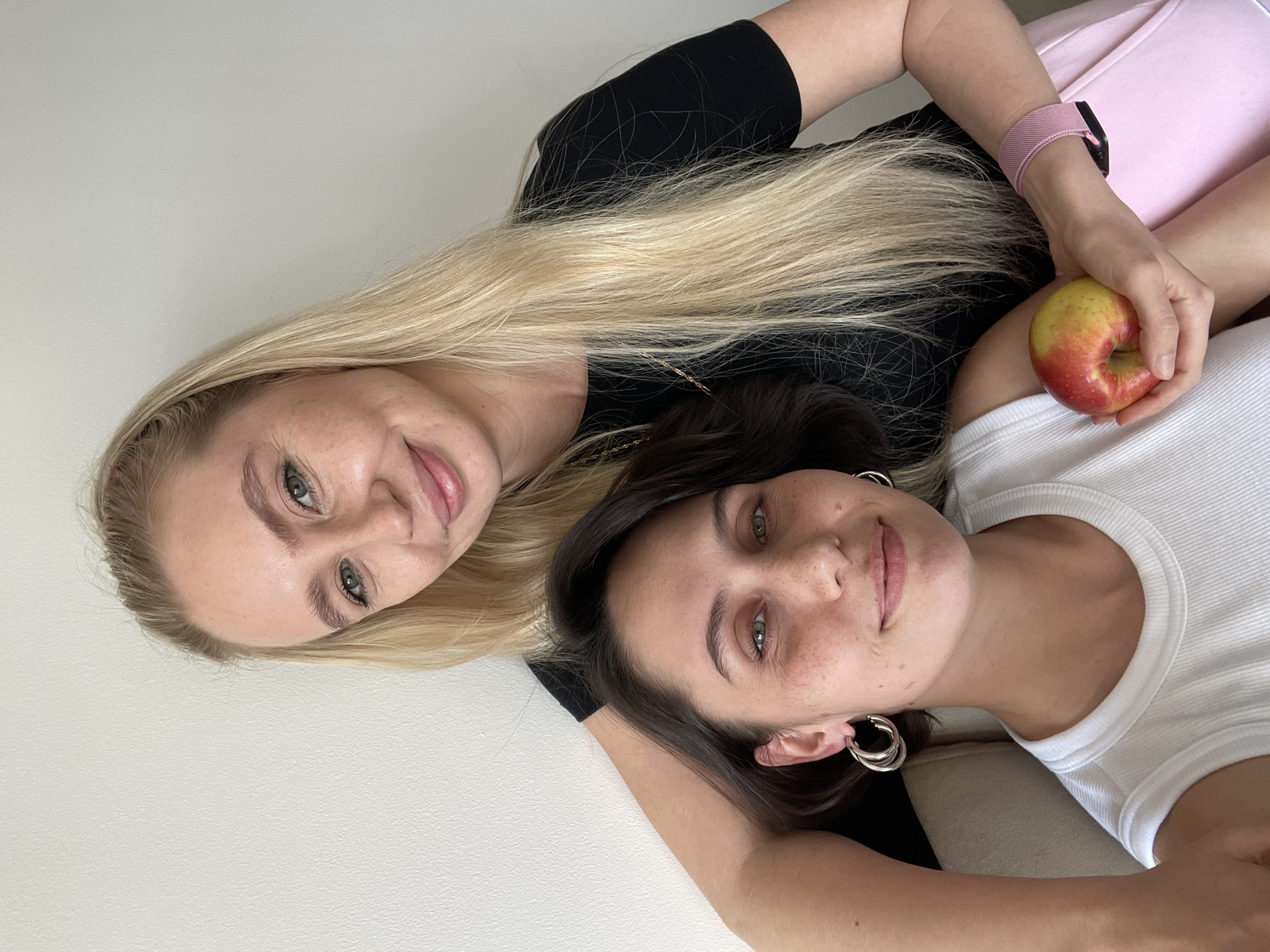 Two women are lying next to each other. The woman on the left has long blonde hair and is wearing a black top. The woman on the right has dark brown hair and is wearing a white ribbed tank top. One of the women is holding an apple. There is a white wall in the background. There is no evidence to suggest this photo was taken in Izhevsk, Russia.