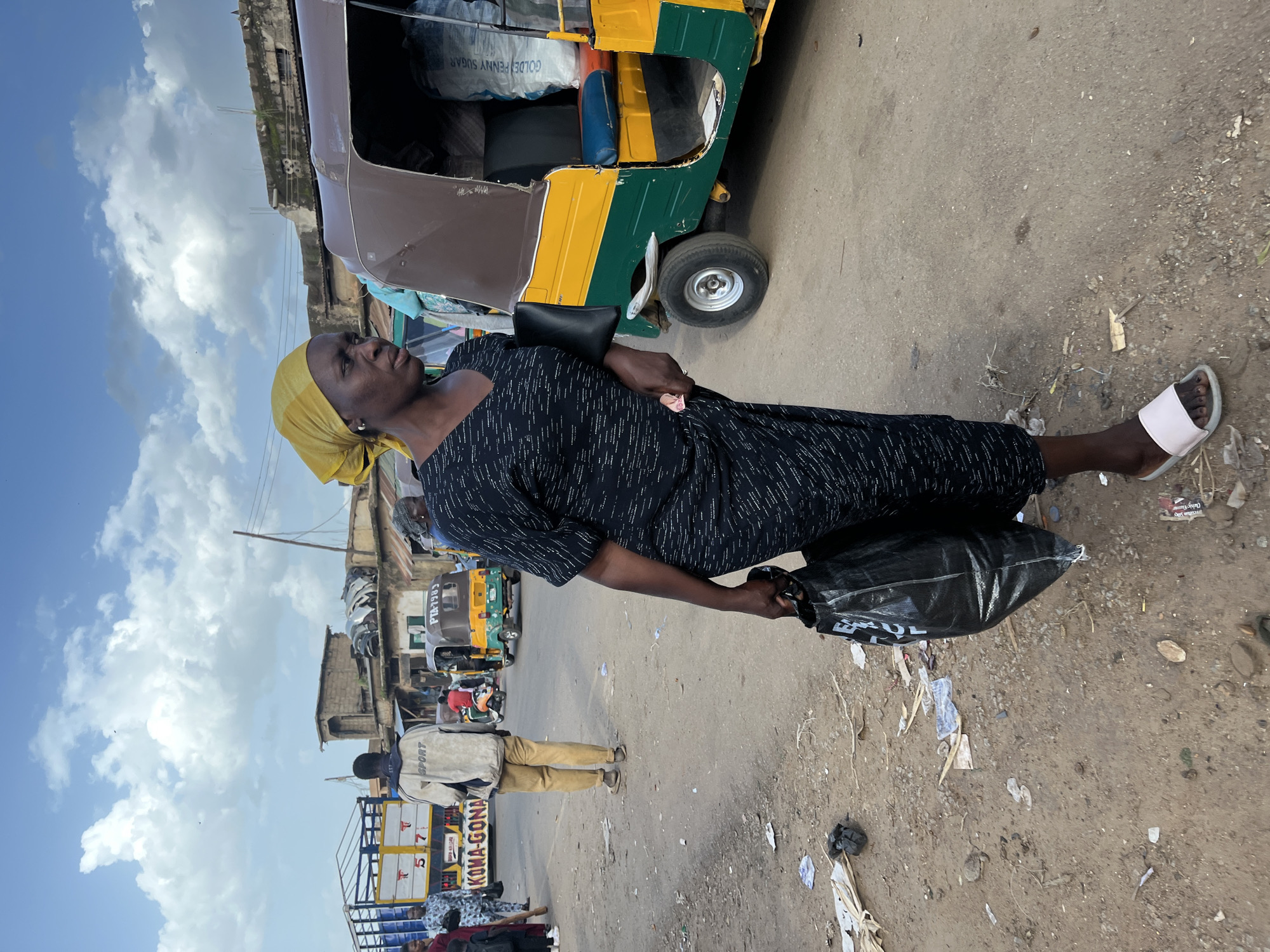 A medium shot captures a woman in a black patterned dress and yellow headscarf walking right to left in an outdoor market scene in Rafin Par, Nigeria. She carries a large black plastic bag in her right hand and a dark clutch in her left. The ground is a dirt and asphalt mixture littered with debris. Behind her, a brightly colored auto-rickshaw, green, yellow, and brown, is parked, with a large bag labeled "GOLDEN PENNY SUGAR" visible inside. Further in the background, a truck with the words "KOMA-GONA" is visible, along with other vehicles and structures indicative of a bustling marketplace. The sky is cloudy with patches of blue.