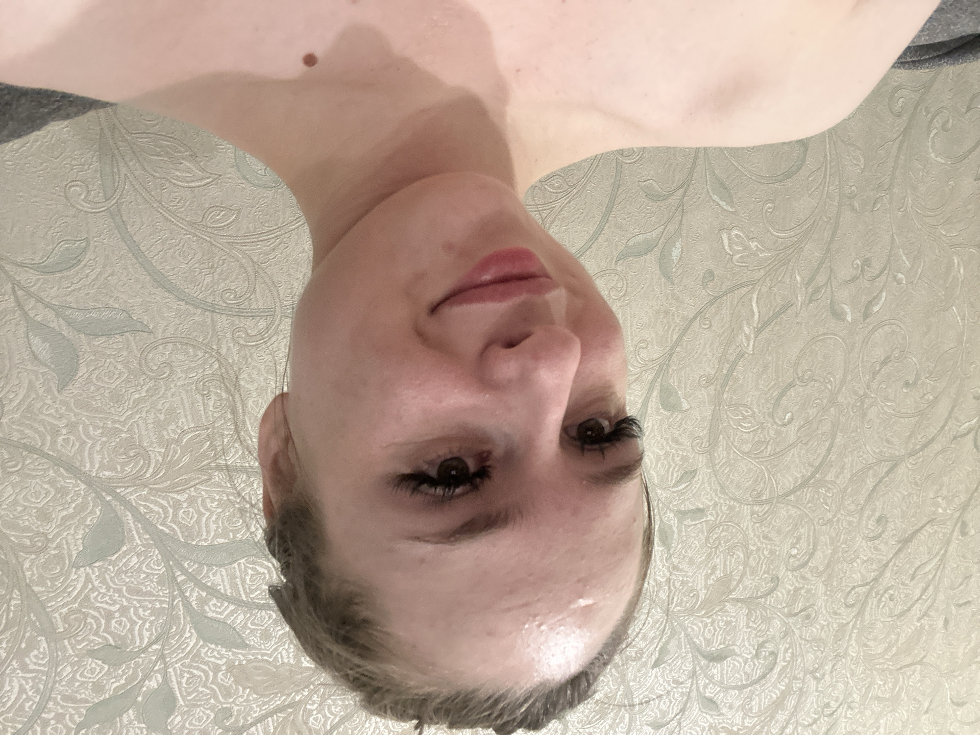 A young woman with light skin, dark eyebrows, and dark eyelashes is pictured from the chest up, facing slightly to her left. Her hair is pulled back, and her forehead appears slightly sweaty. She is wearing no visible clothing on her upper body. The background is a textured wallpaper with a light, ornate pattern of swirling vines and leaves in muted green and cream tones. To the lower left of the frame, a dark grey fabric is visible, possibly part of a chair or sofa. The lighting is soft and appears to be coming from the front.