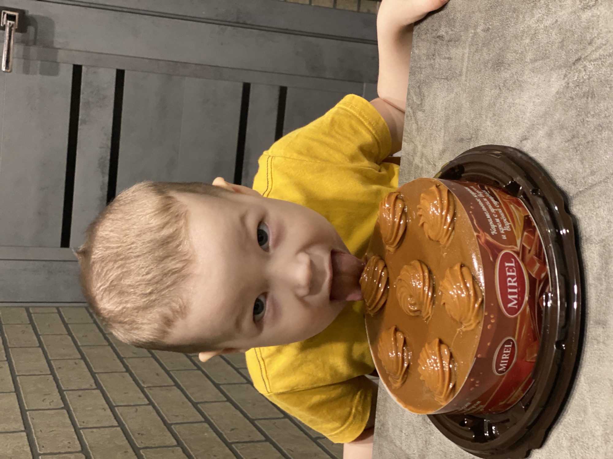 A young child in a yellow shirt is licking the top of a "Mirel" brand cake. The cake is round and covered in a caramel-colored frosting with decorative swirls. The background consists of a brick wall and a gray wall, suggesting an indoor setting, possibly a kitchen. There is no evidence indicating this photo was taken in Sochi, Russia.