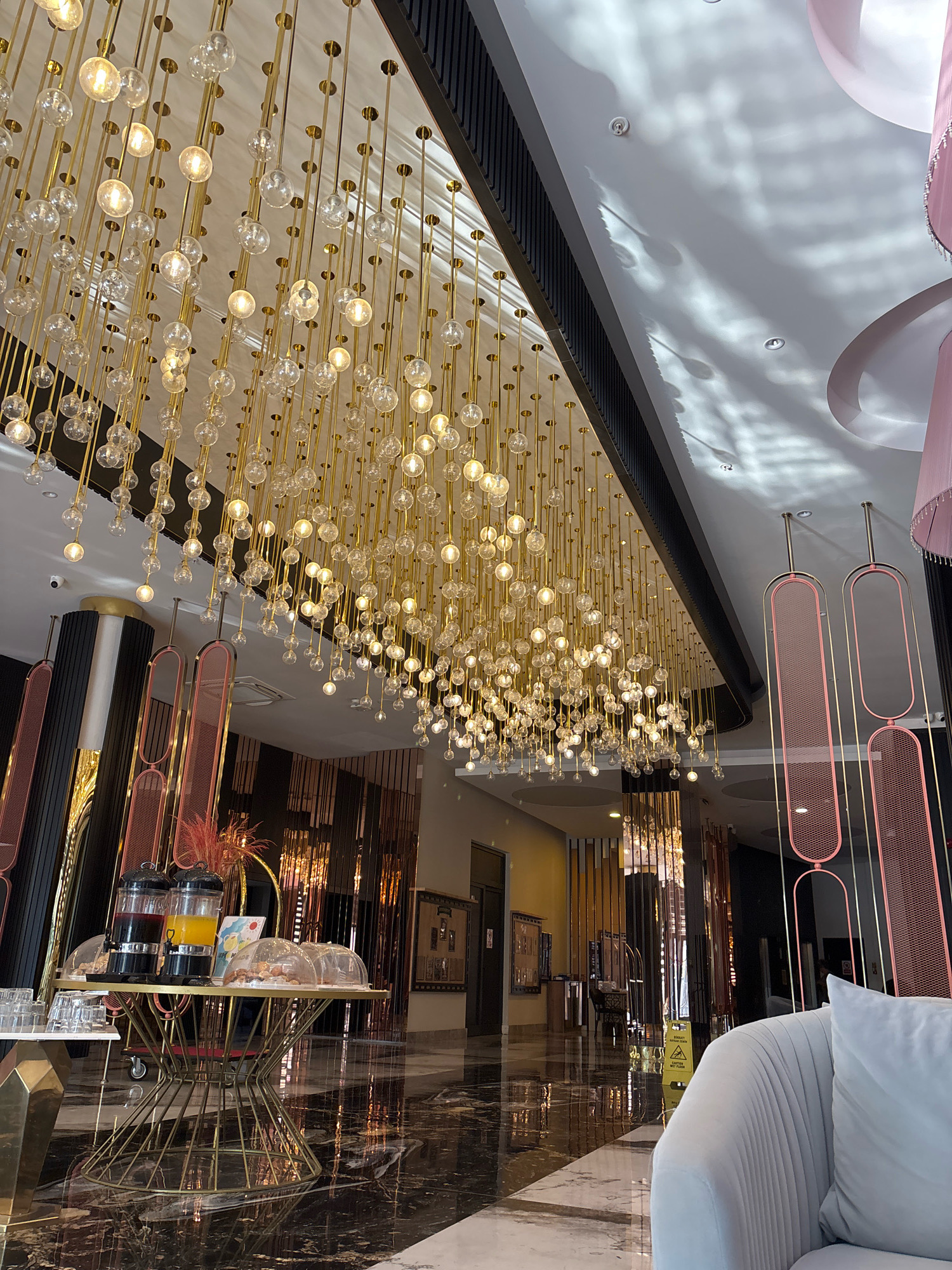 The image depicts the luxurious interior of a hotel lobby, likely in Alanya, Turkey, characterized by its opulent design and extensive use of gold accents. Dominating the scene is a grand, cascading chandelier composed of numerous clear glass spheres suspended by thin gold rods, illuminated to cast a warm glow and create a shimmering effect on the ceiling and surrounding surfaces. The ceiling itself is white and features intricate, almost ethereal shadows cast by the chandelier's lighting, along with discreet recessed lighting fixtures and what appear to be fire safety devices.
In the mid-ground, a decorative buffet table with a gold geometric base holds two dispensers filled with dark red and orange beverages, respectively. Beside them are covered trays containing pastries or other food items, and a stack of clear drinking glasses. The table is adorned with a small, colorful card. Behind the table, the lobby features polished dark marble flooring that reflects the overhead lighting and architectural details, contributing to the overall sense of grandeur.
The background reveals further elements of the hotel's sophisticated decor, including tall, striped pillars with gold detailing, and decorative screens or dividers with pink mesh and gold frames. The overall ambiance suggests a high-end establishment designed for comfort and aesthetic appeal, likely catering to tourists in a popular resort destination like Alanya. There are no visible people in the frame, and the scene appears to be captured during daylight hours, evidenced by the natural light subtly mixing with the artificial lighting. The focus is on the architectural features, lighting, and decorative elements of the lobby.