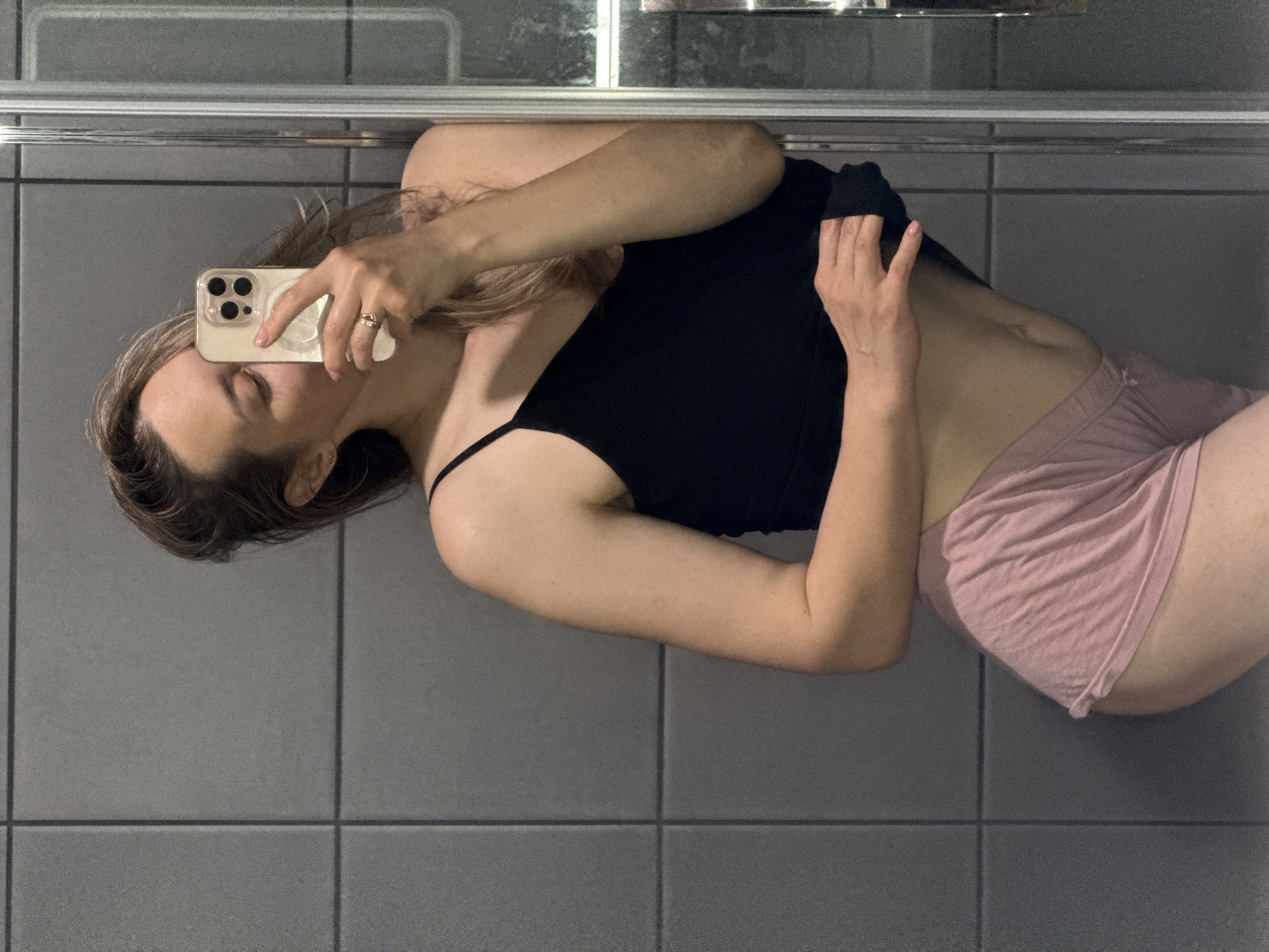 This rotated mirror selfie captures a person, likely female, lying or leaning against a tiled surface in an indoor setting. The individual has light brown or blonde hair and is dressed in a black spaghetti strap top and light pink or peach shorts/underwear. They are holding a light-colored smartphone with a clear case and a distinct three-lens camera module, obscuring most of their face as they take the picture. A ring is visible on their right hand.
The setting appears to be a bathroom or a similar utility space, characterized by uniform dark gray square tiles covering the wall and floor. A horizontal metallic bar or frame, possibly part of a mirror or shower enclosure, runs across the upper portion of the image, reflecting some light. The lighting is soft and appears to be artificial indoor illumination, creating a somewhat dim ambiance. The person's eyes are closed, suggesting a relaxed pose or concentration on the phone's screen. No text is visible in the image. While the location context is Chaikovsky, Russia, the visual details in the image do not provide specific cues to pinpoint an exact location within the city.