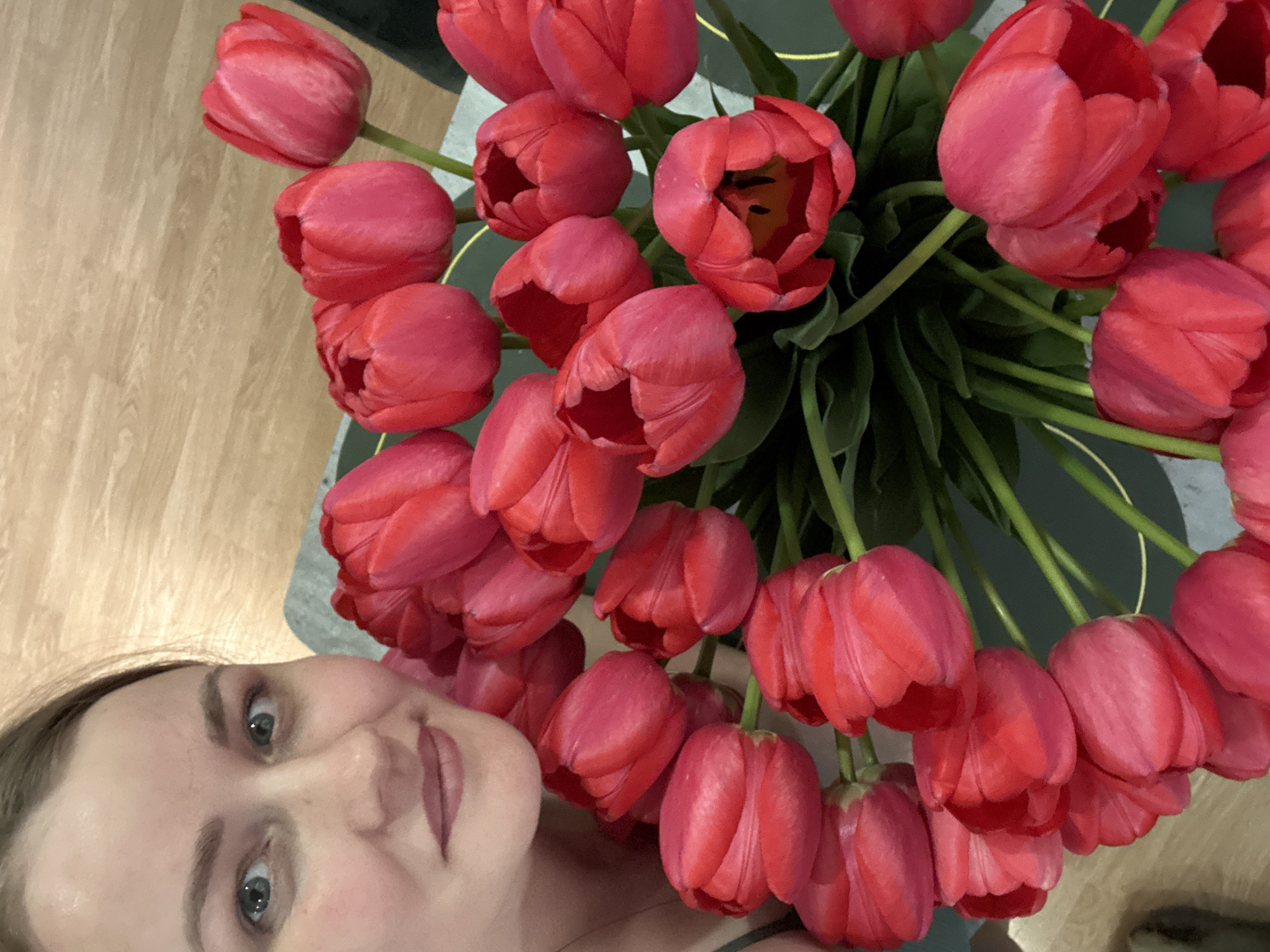 A person with light skin, light brown hair, and blue eyes is shown from the nose up on the left-hand side of the frame. The person's face is partially obscured by a large bouquet of red tulips that take up the majority of the frame. The tulips are arranged in a circular shape, with green stems and leaves visible between the flower heads. The photo was possibly taken in the city of Chaikovsky, Russia, as this information was provided. The setting is possibly indoors.