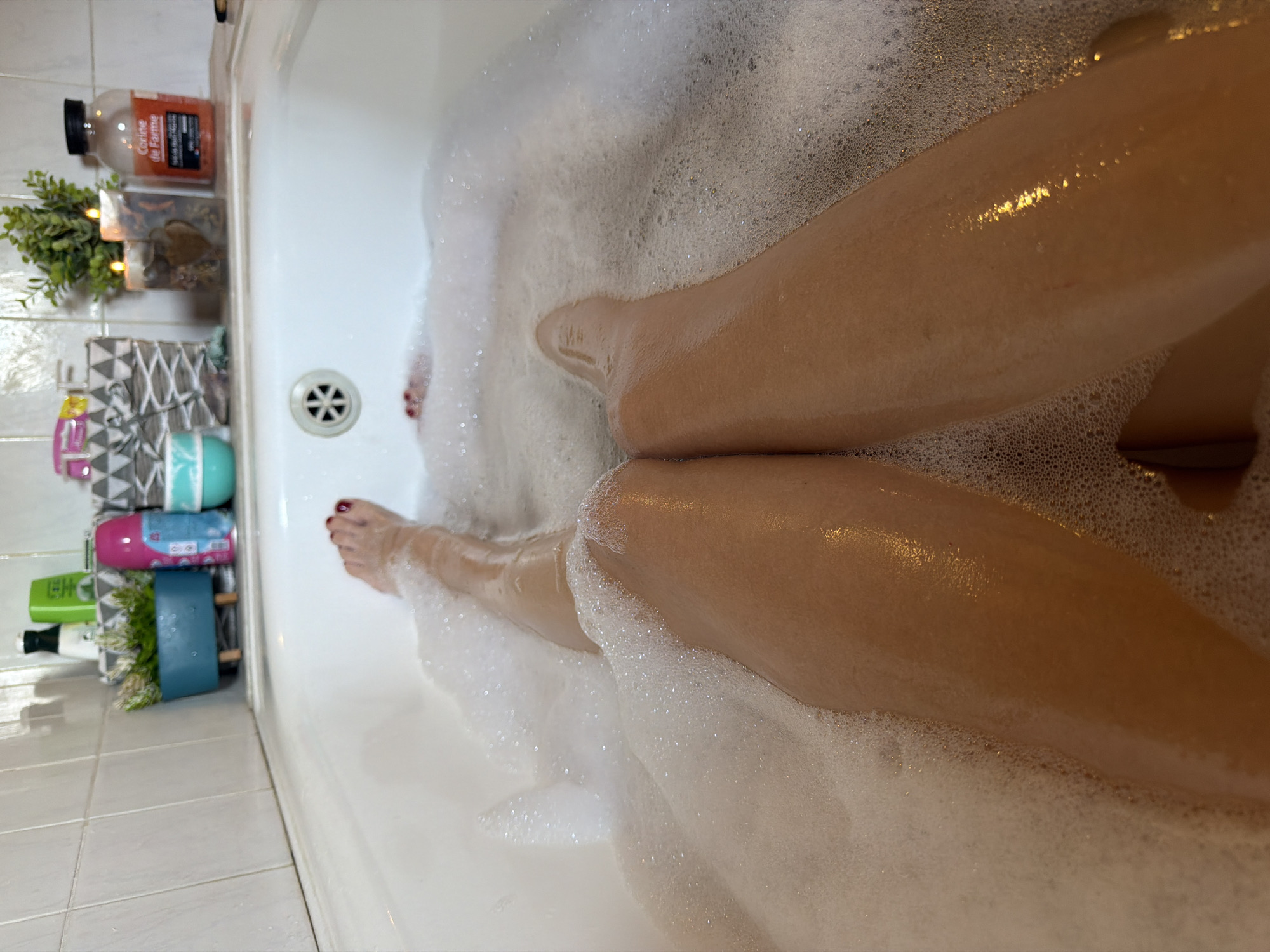 The image shows a person's legs submerged in a bubbly bathtub. The scene is likely a bathroom, as indicated by the tiled wall and the bathtub itself. Several bath products are visible on a shelf next to the tub, including bottles of what appear to be shampoo, conditioner, or body wash, as well as some decorative items. The lighting suggests it could be daytime or a well-lit bathroom. The mood conveyed is one of relaxation and self-care, given the context of a bubble bath. No specific time of day or weather conditions are discernible. There is visible text on one of the bottles, which reads "Caudalie Vinotherapie".